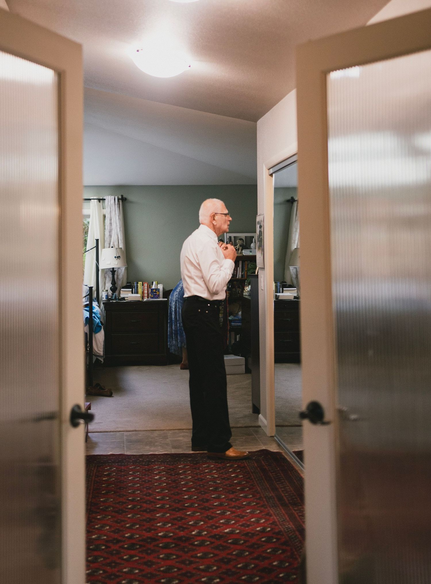 Elderly man in white shirt and black pants getting dressed in a bedroom, viewed through French doors.