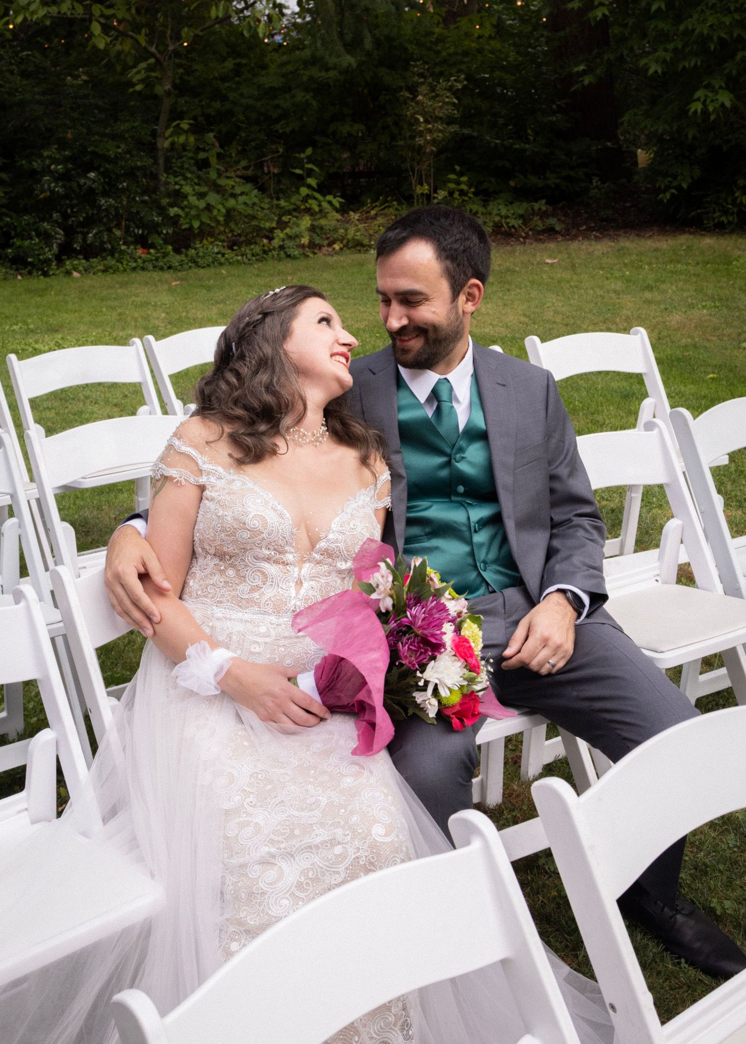 Bride and groom sitting together in white chairs smiling at each other during outdoor wedding ceremony.
