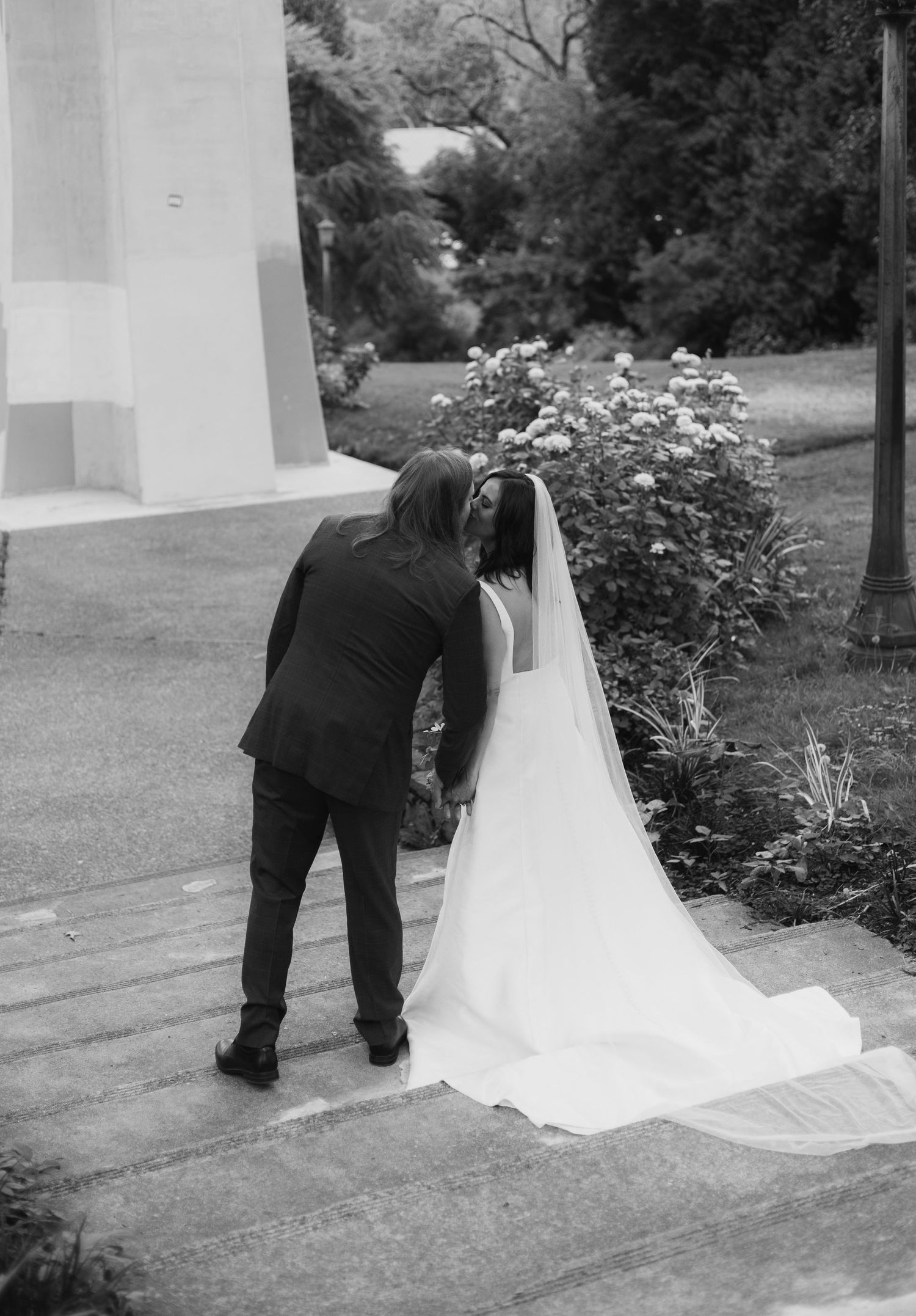 Groom leaning to kiss bride in white wedding gown and veil outdoors.