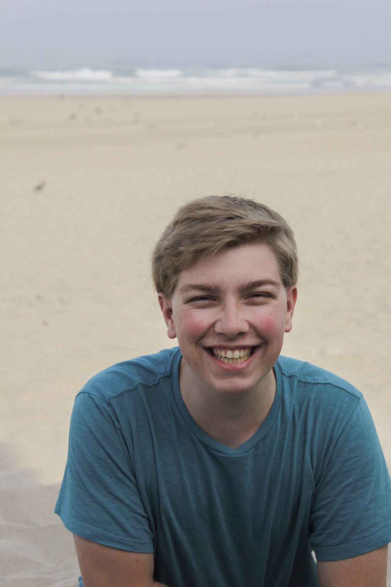 A cheerful teenage boy with short blonde hair laughs on a sandy beach wearing a teal t-shirt.