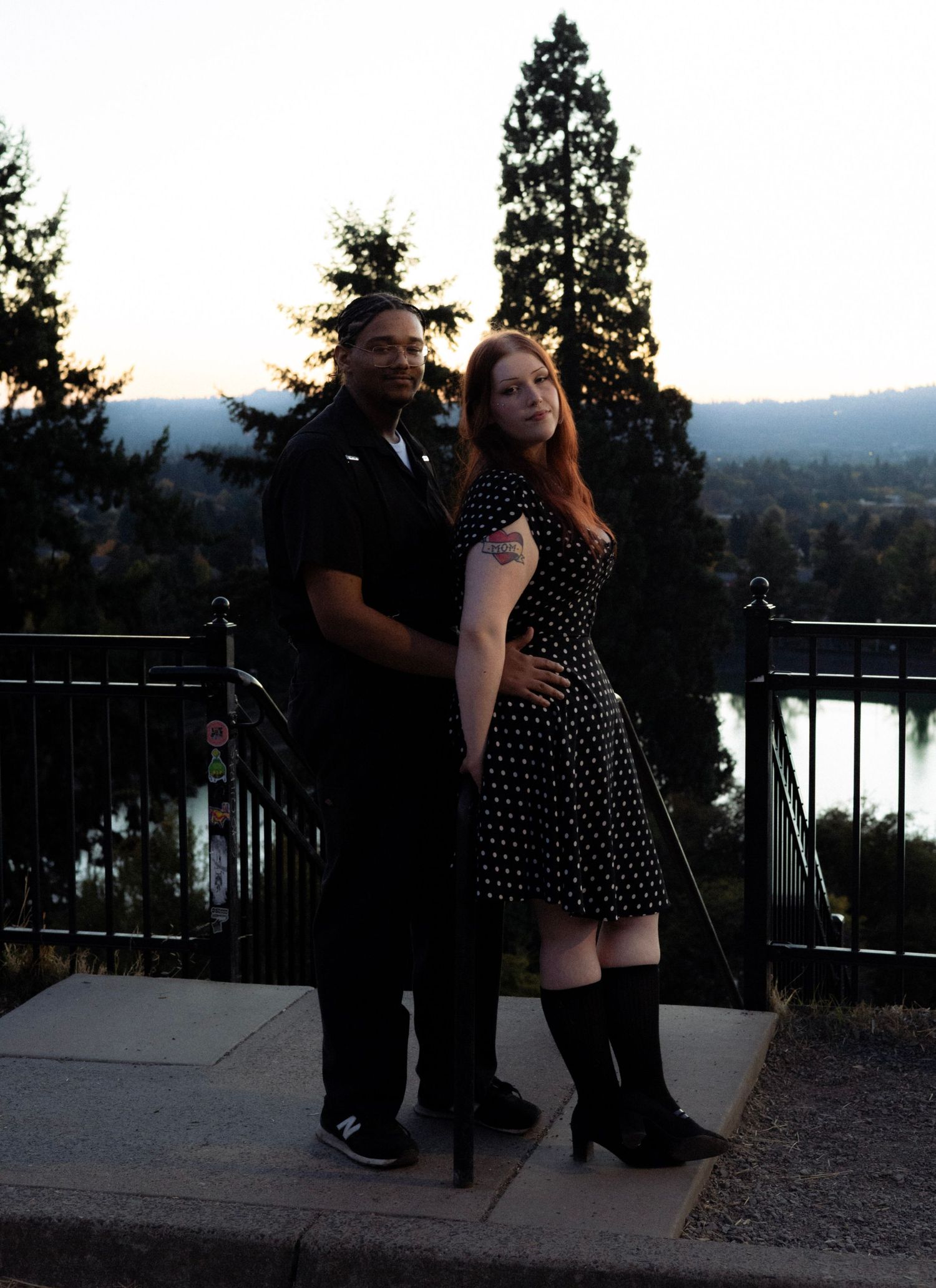 Couple in formal attire posing at scenic overlook with river and trees at dusk, woman in polka dot dress and boots.