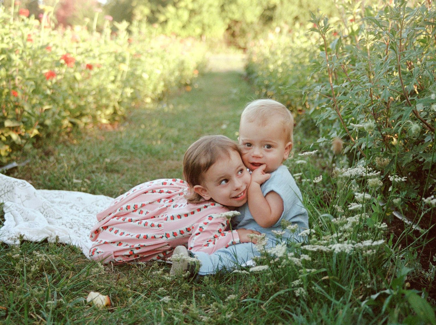 Two young siblings cuddle together on a white blanket in a lush green garden with colorful flowers in the background.