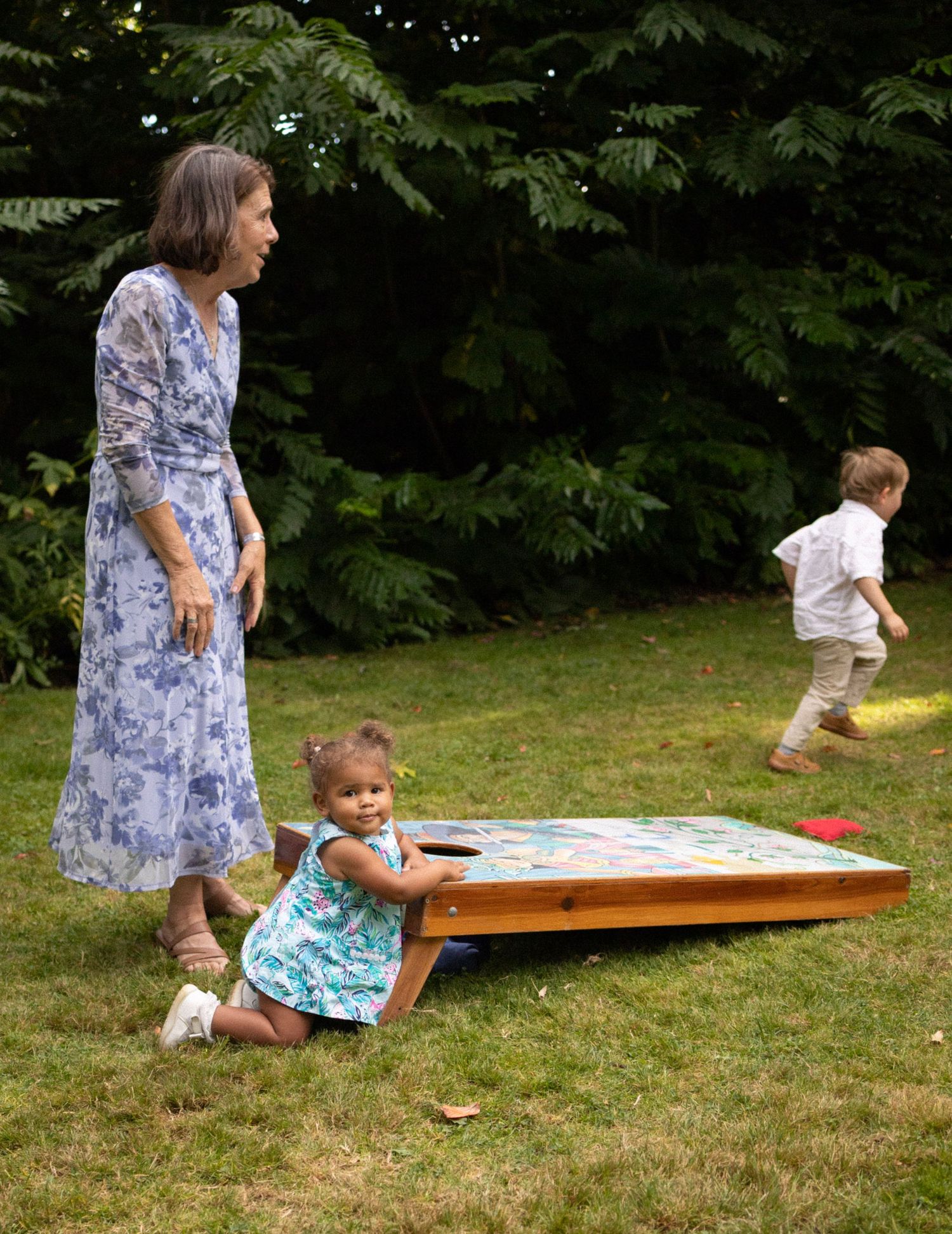A woman in a blue floral dress watches a toddler girl play cornhole on a sunny lawn at an outdoor gathering.