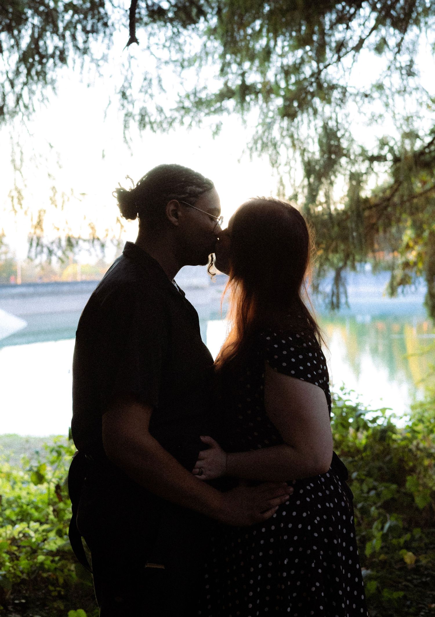 Couple sharing a kiss in silhouette outdoors, woman in polka dot dress with baby bump, green trees and water in background.