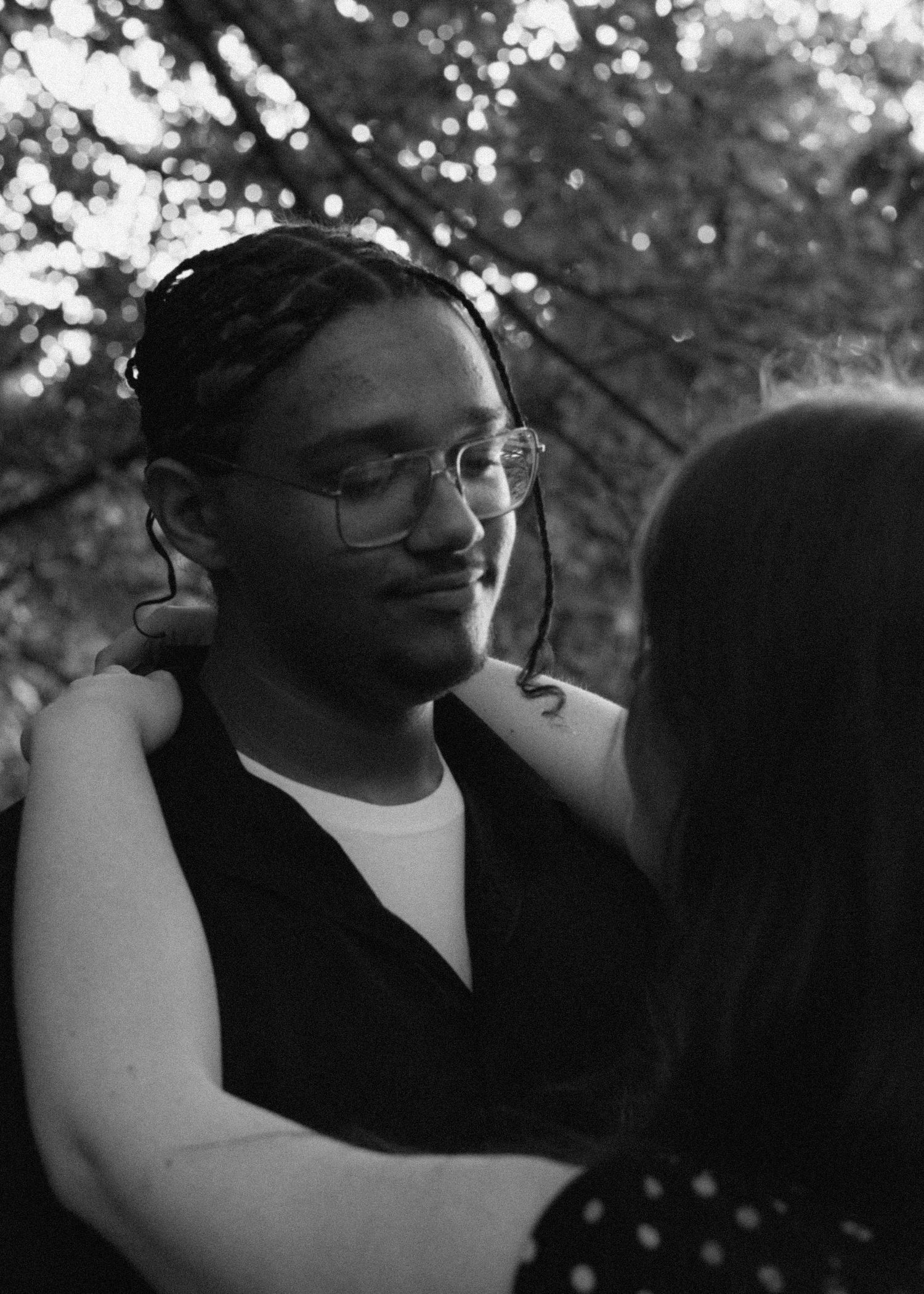 Black and white photo of a man with glasses and braids embracing a woman outdoors under trees.