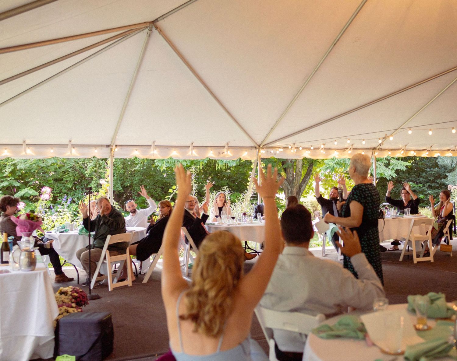 Guests celebrate under a white tent at an outdoor wedding reception with string lights and lush greenery.