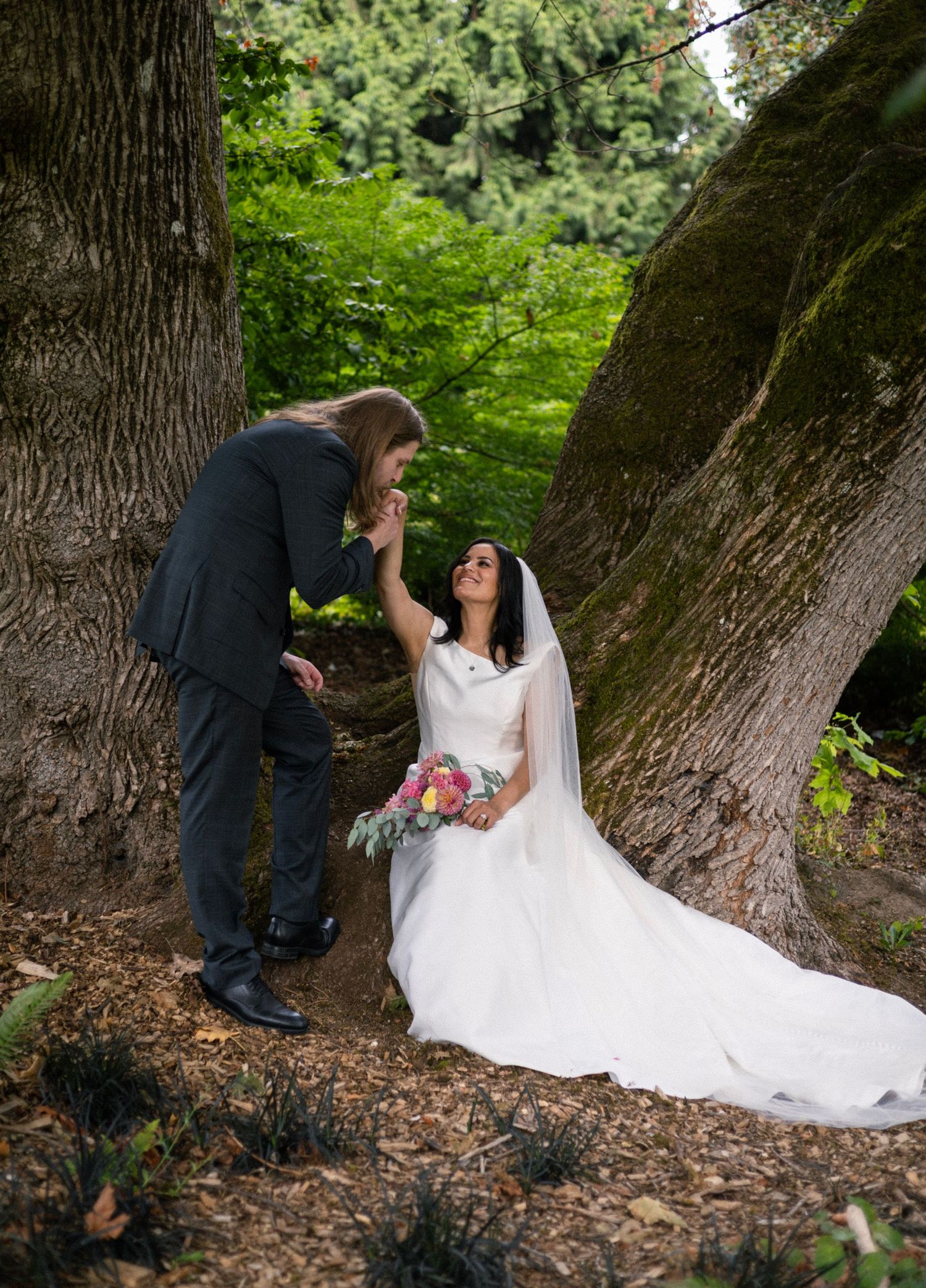 Groom kisses bride's hand as she sits in white gown with bouquet between two large moss-covered trees in lush garden.