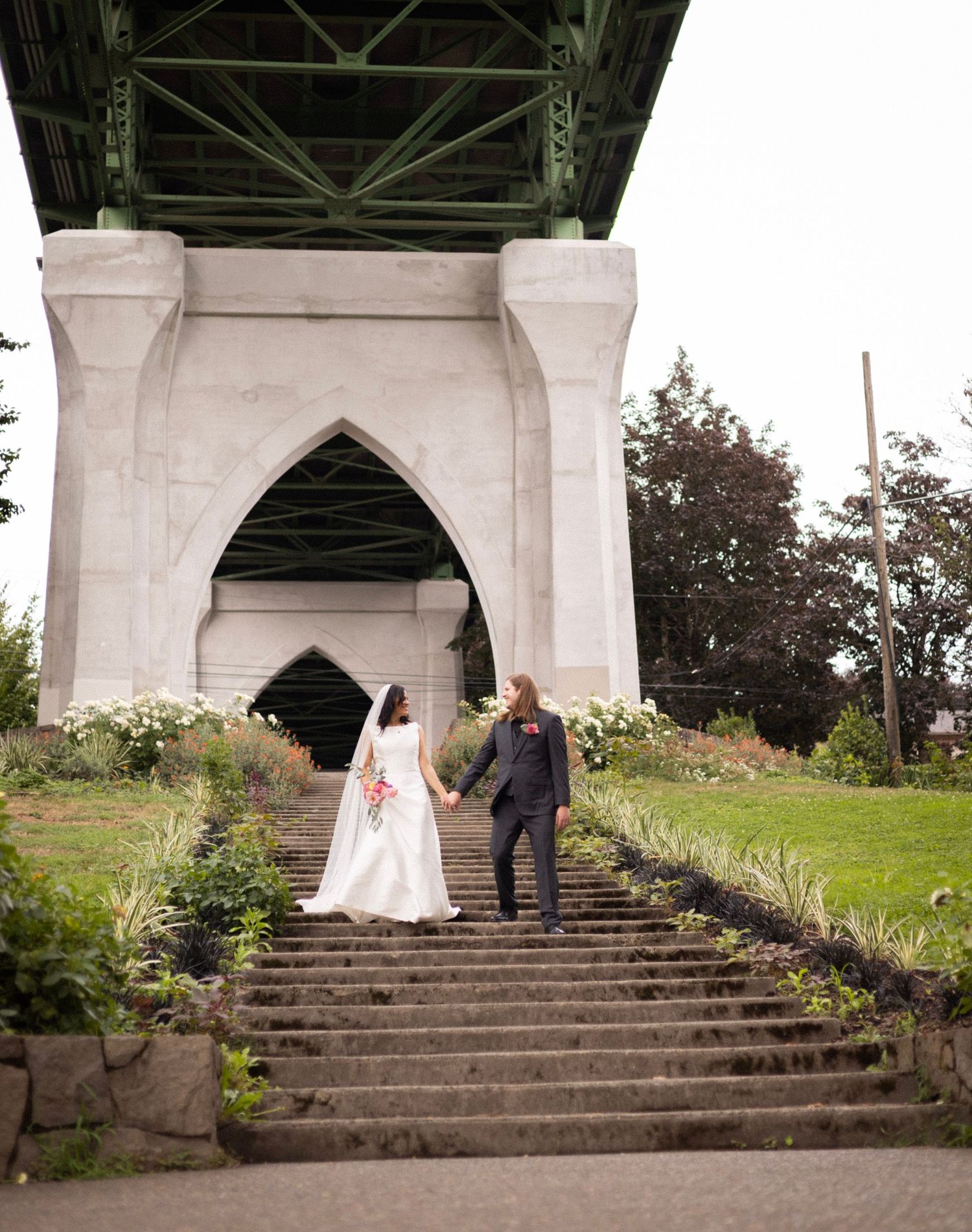 Bride and groom hold hands on stone steps beneath a grand bridge arch, surrounded by lush greenery on their wedding day.