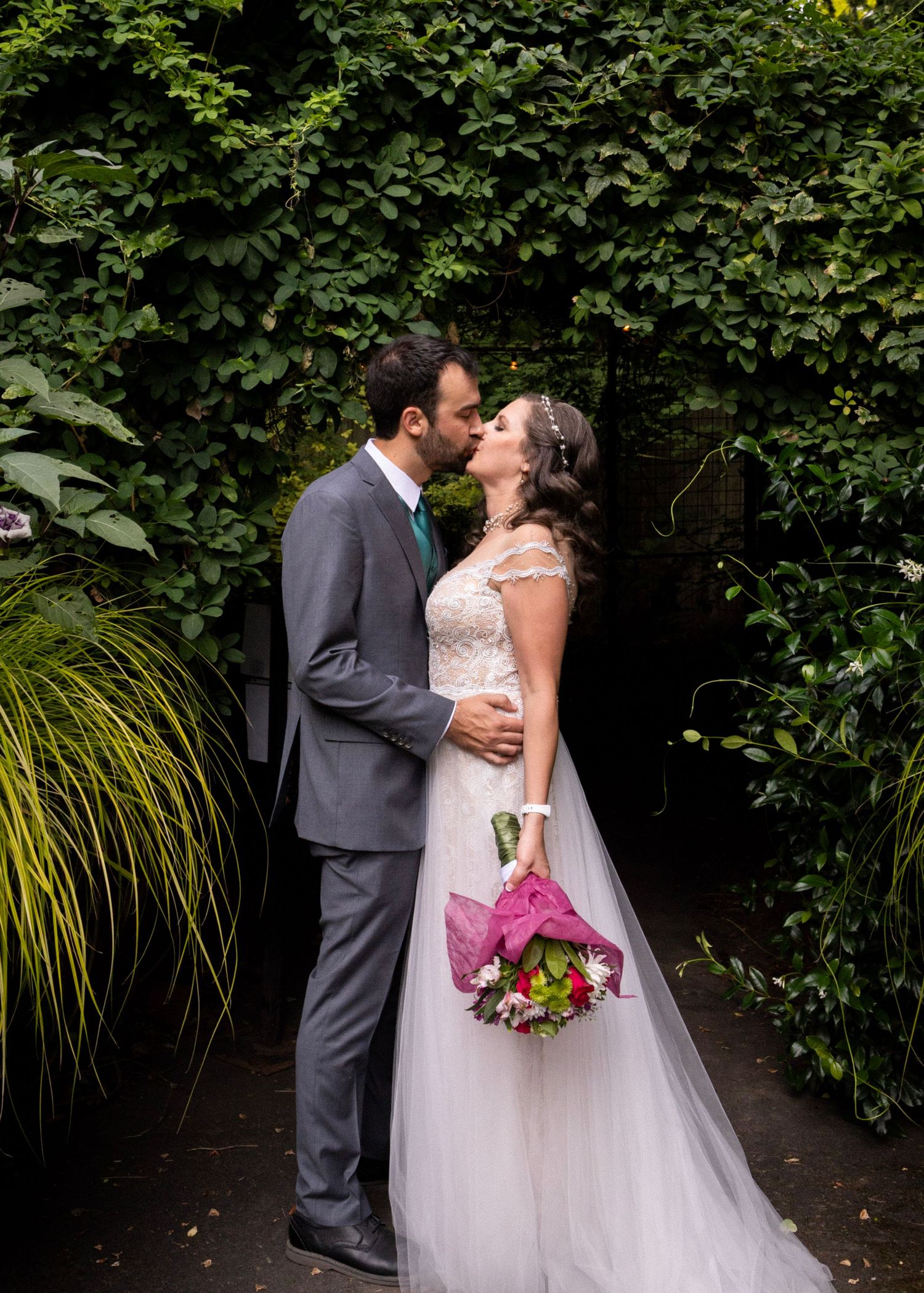 A bride and groom share a romantic kiss surrounded by lush green ivy at their outdoor wedding.