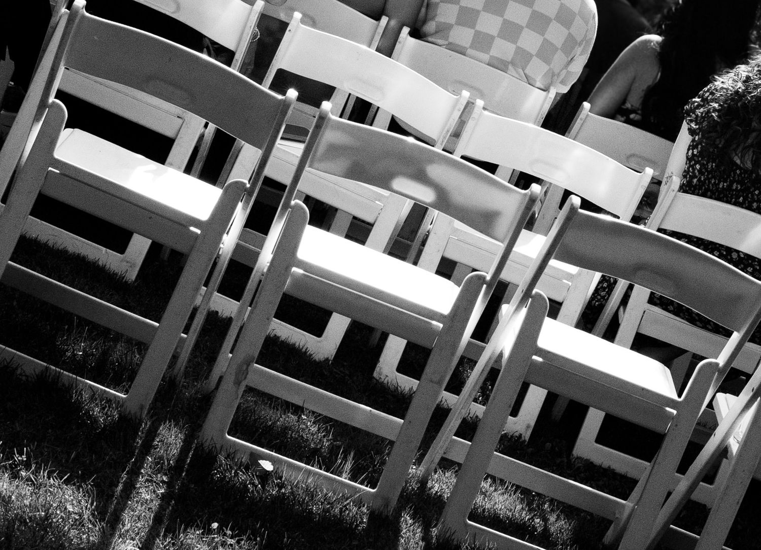 Rows of white wooden folding chairs arranged outdoors on grass for a wedding ceremony in black and white.