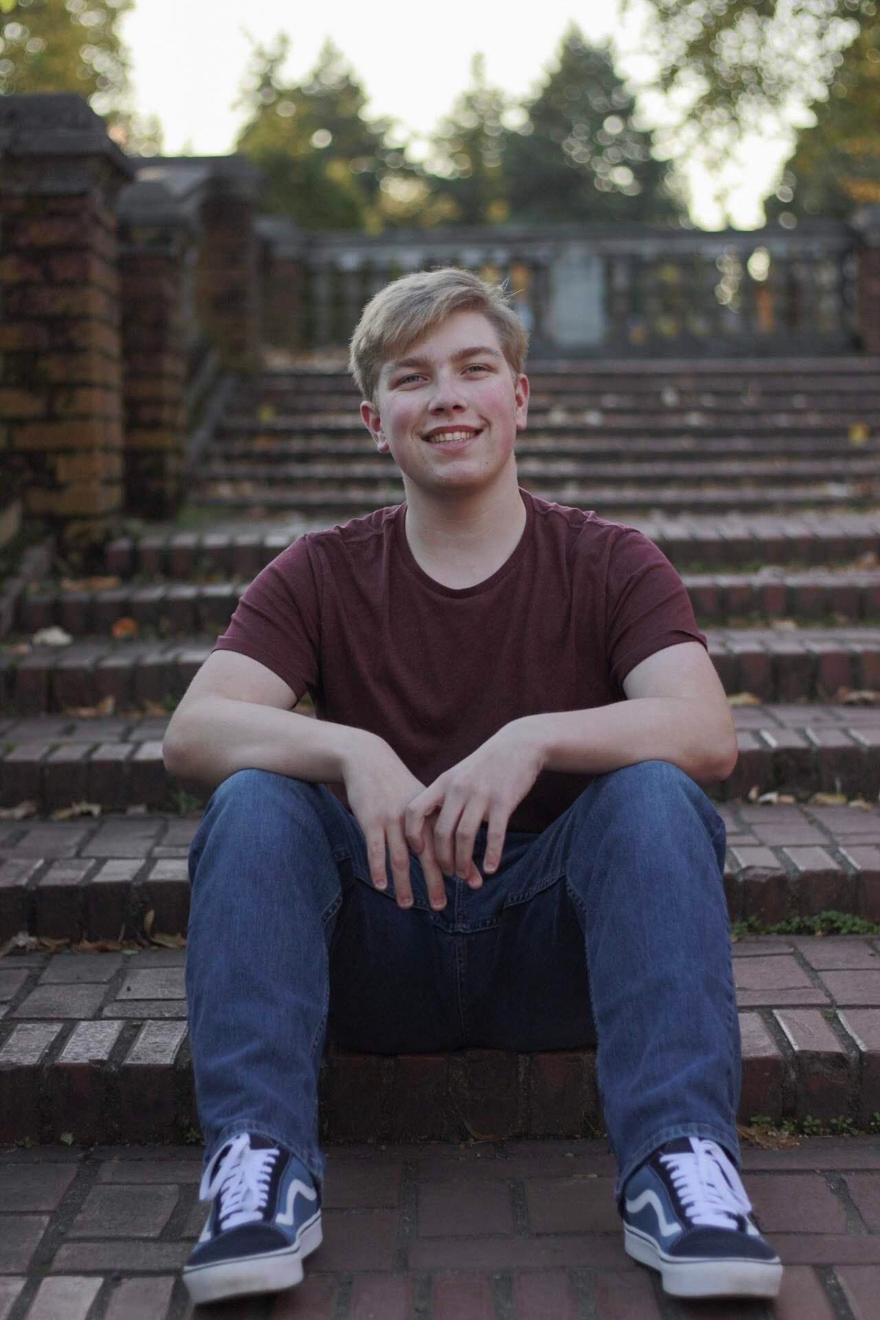 Smiling teenage boy with blonde hair sits on brick steps wearing maroon shirt and jeans.