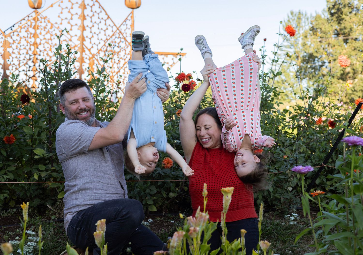 Happy family with two parents swinging their young children upside down in a colorful flower garden.