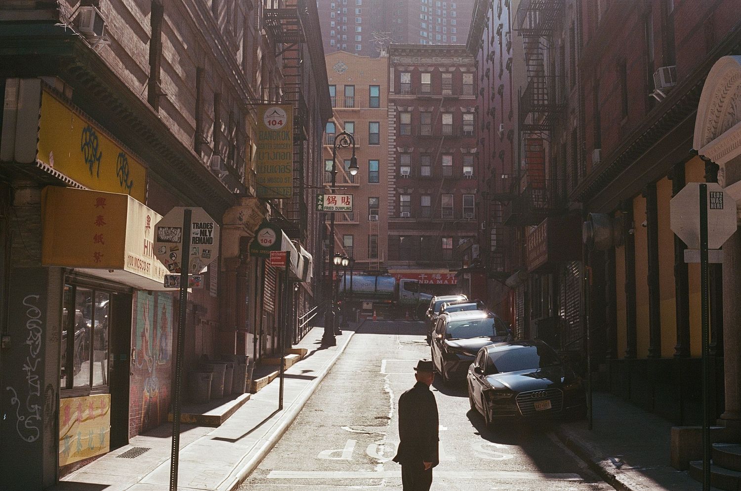 Sunlit narrow alley in New York City with parked cars and lone figure.