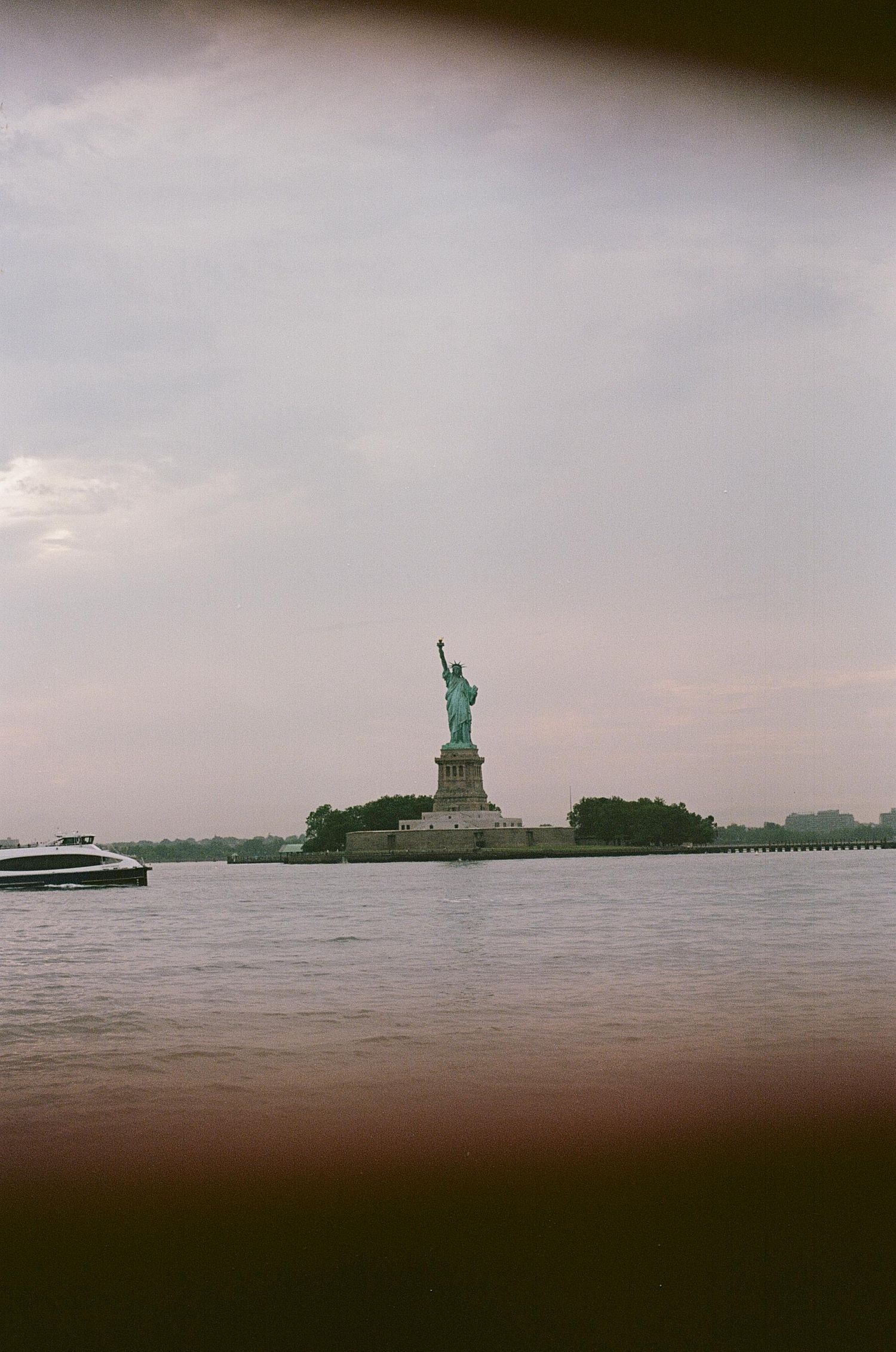The Statue of Liberty stands on Liberty Island in New York Harbor with a boat passing by under cloudy skies.
