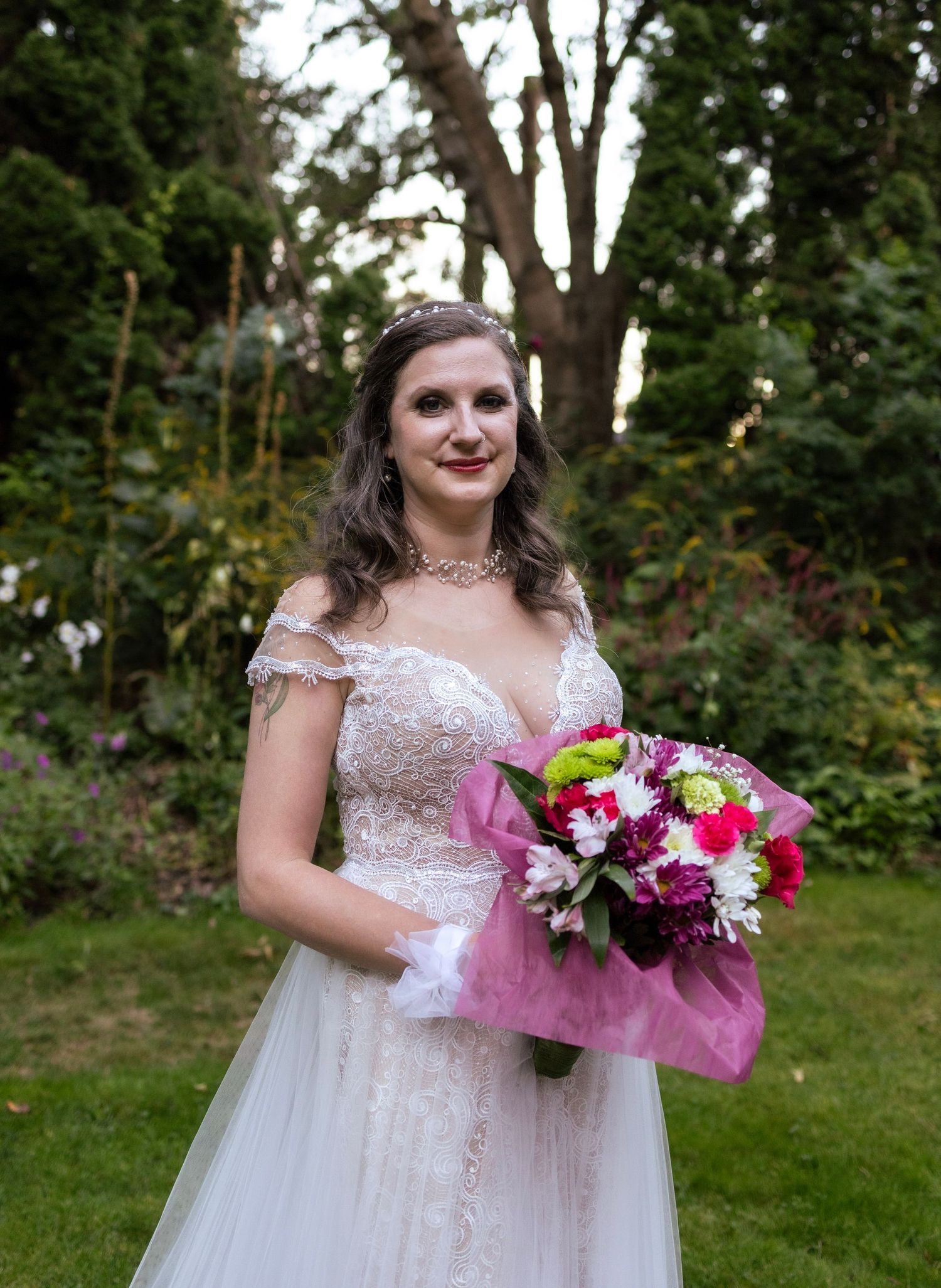Bride in lace wedding gown holding colorful bouquet wrapped in pink, standing in garden with trees.