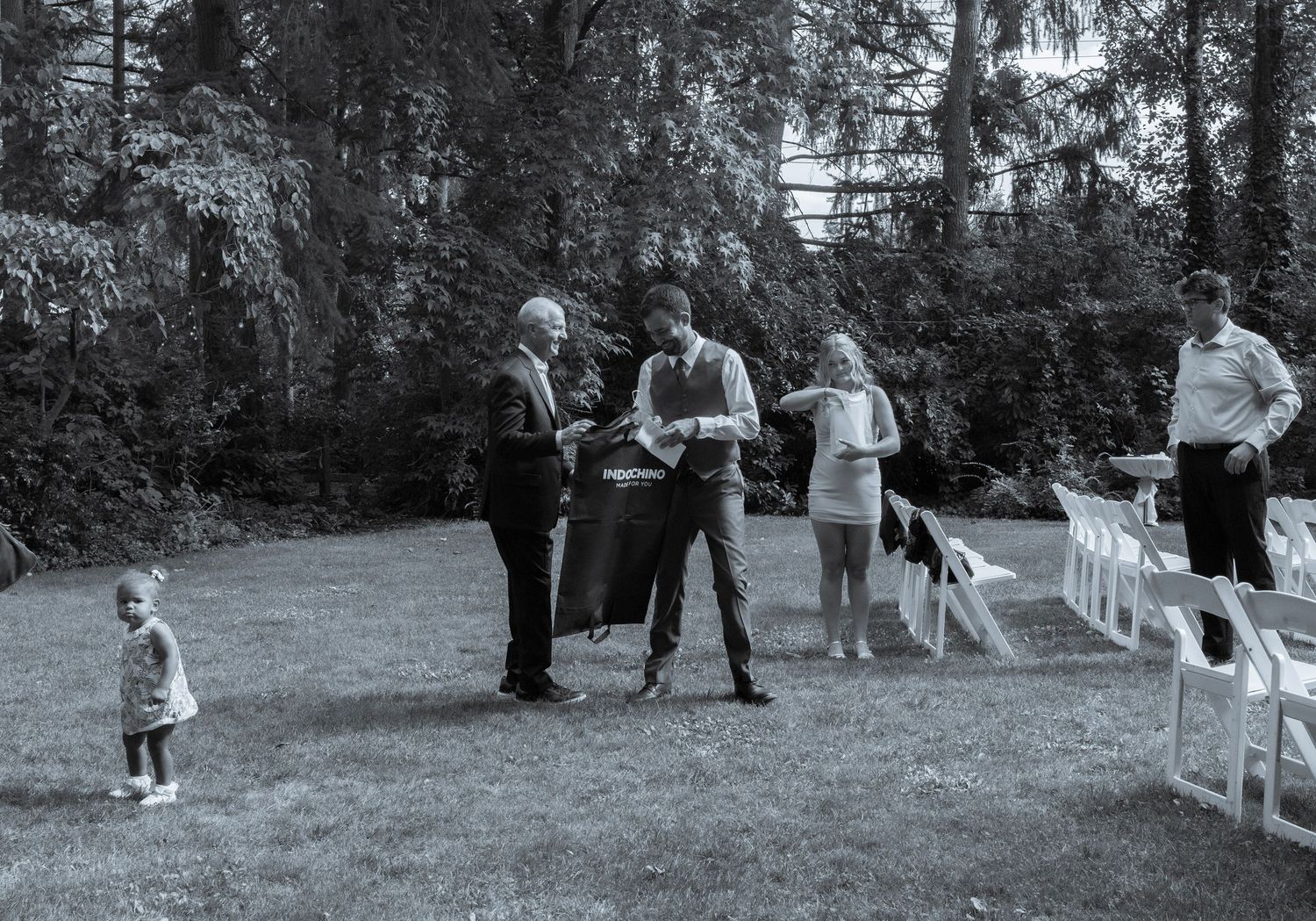 Wedding guests mingle on lawn near white chairs in black and white outdoor photo.