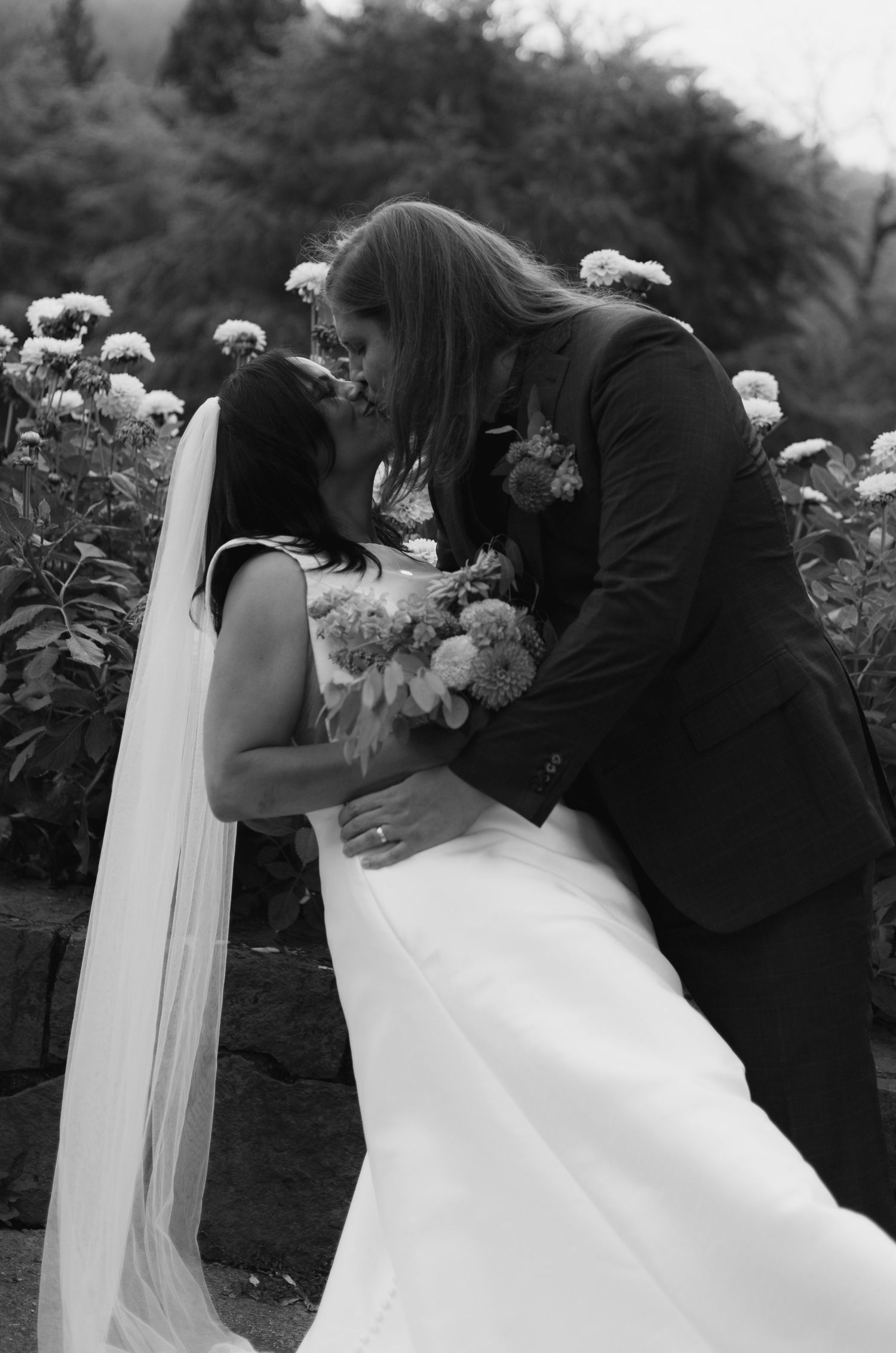 Black and white photo of a bride and groom sharing a romantic kiss in a flower garden on their wedding day.