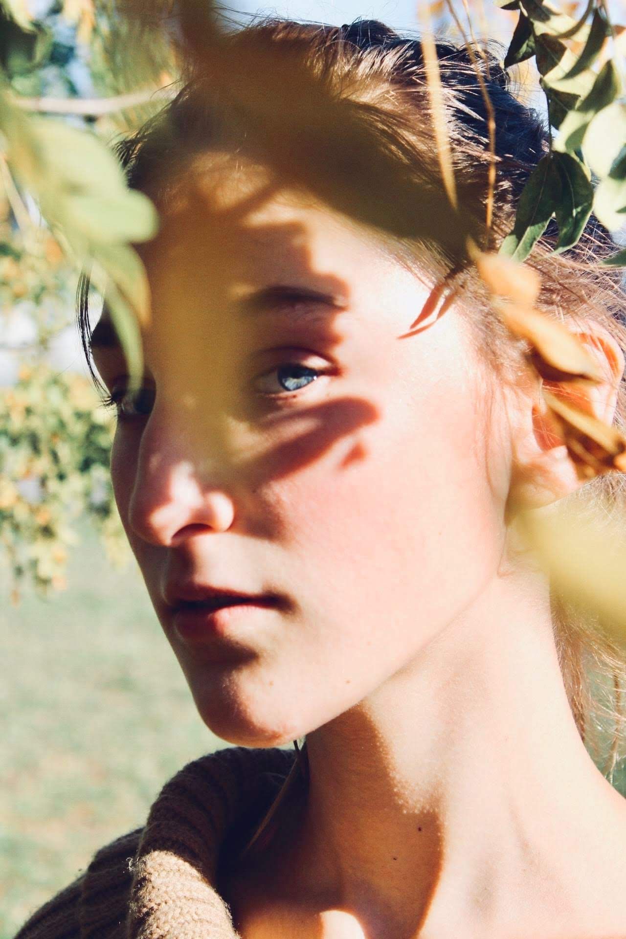 A young woman with dappled sunlight and leaf shadows across her face outdoors.