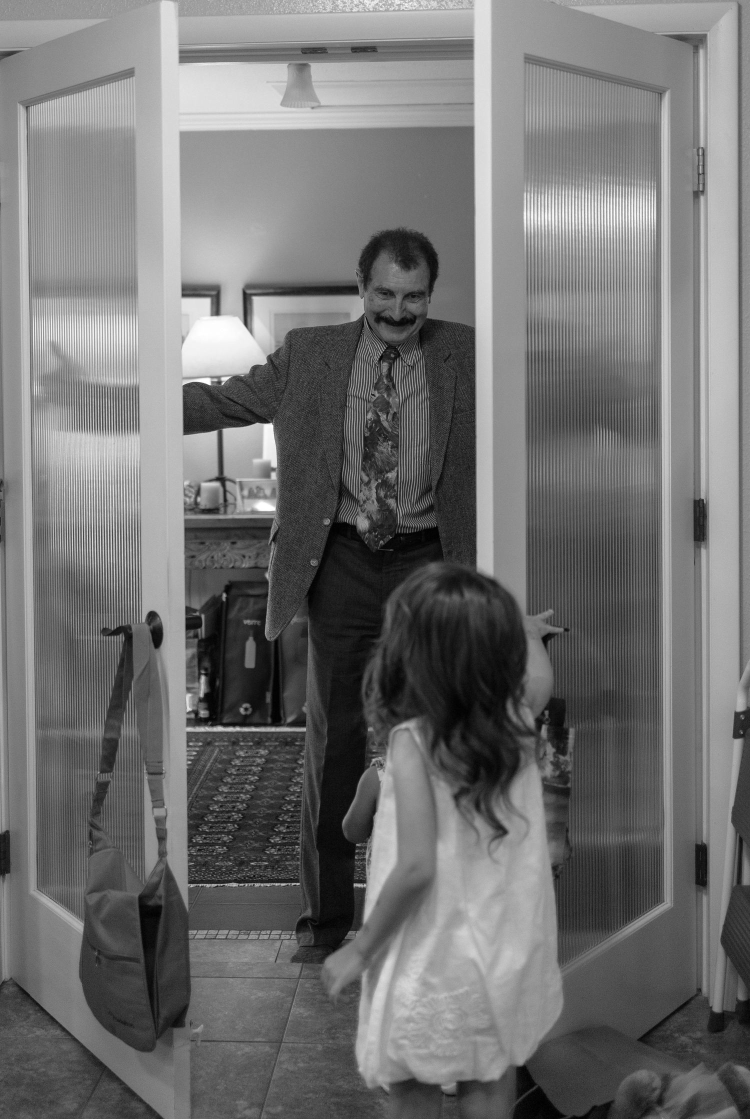 Black and white photo of a man in a suit opening French doors as a young girl in white dress runs to greet him.