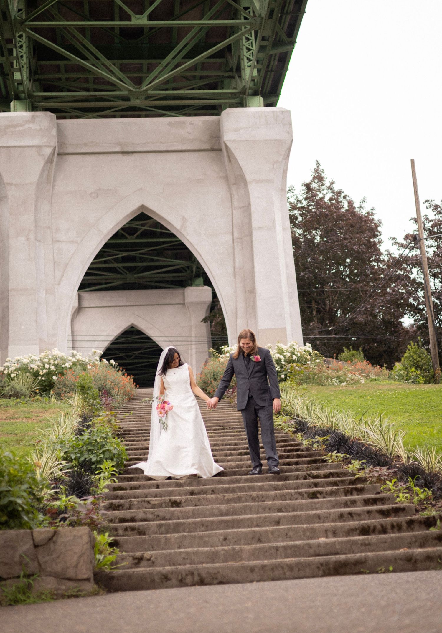 Bride and groom holding hands walking down stone steps under a bridge.