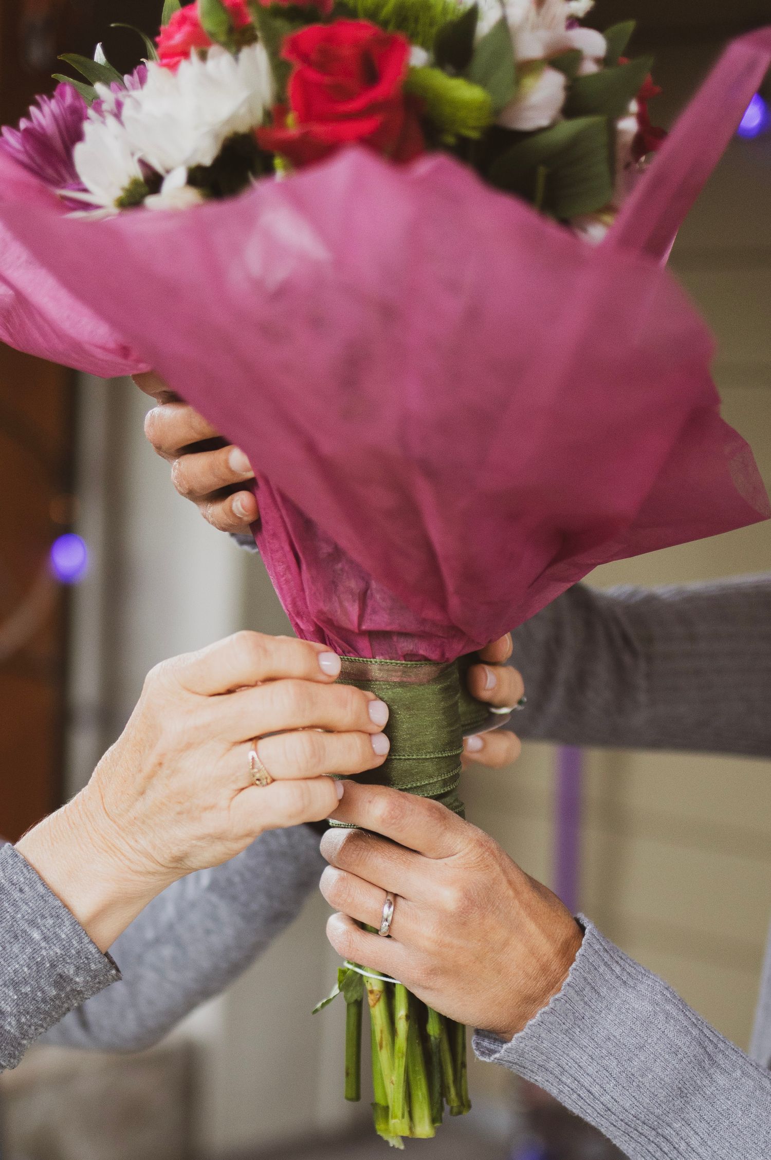 Two people holding floral bouquet wrapped in pink tissue, both wearing wedding rings.