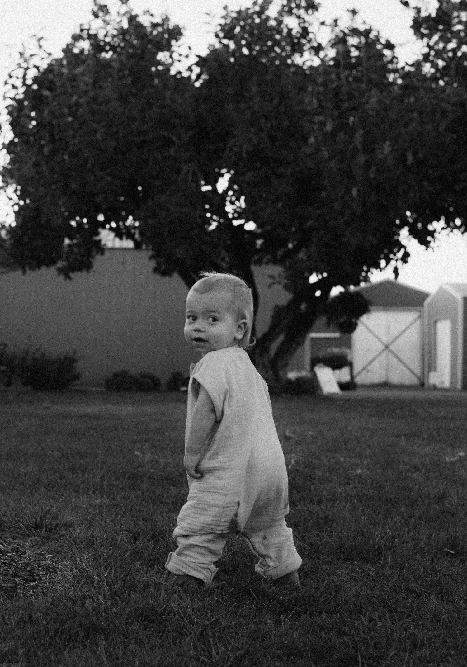 Black and white photo of a toddler in a romper standing on grass with trees and a shed in the background.