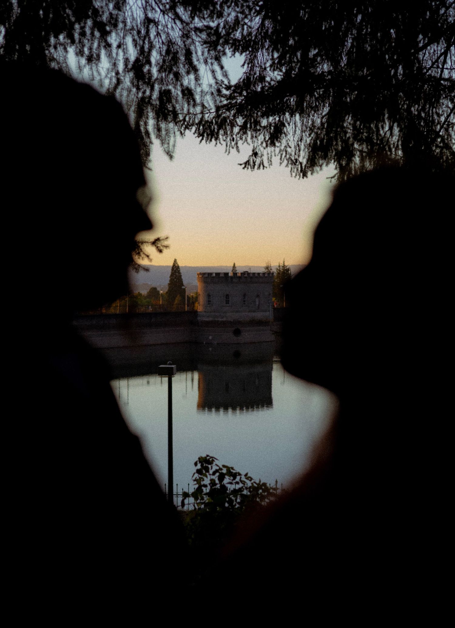 Silhouettes of two people facing each other framing a reflective lake and historic castle at golden hour sunset.