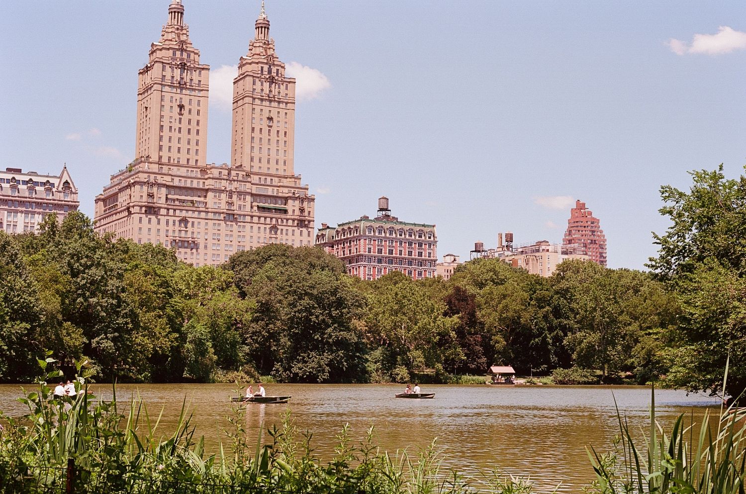 View of tall museums and architecture past lush greens of central park, with people in rowboats on the water nearby.