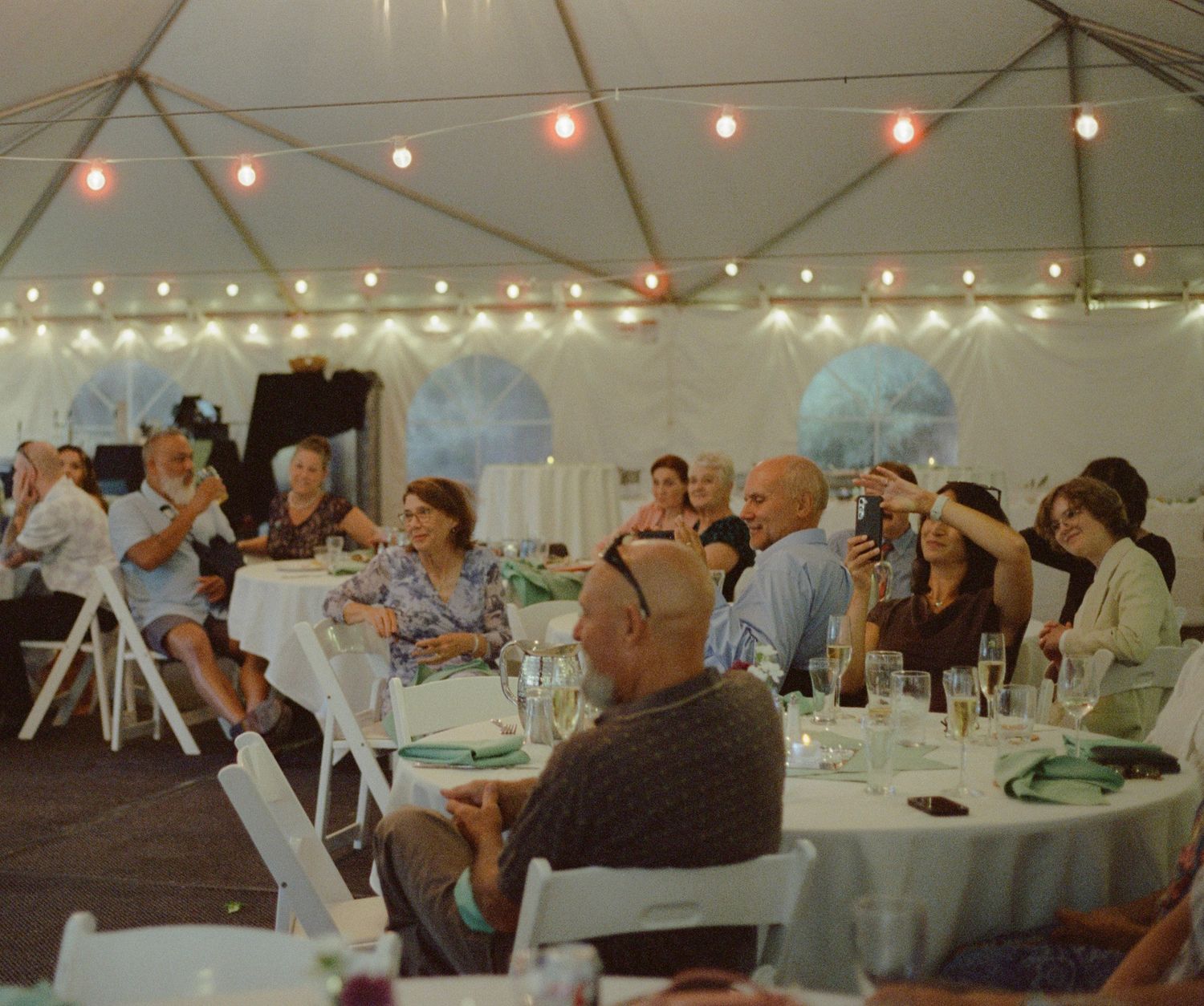 Guests seated at round tables under a white tent with string lights at an elegant outdoor event.