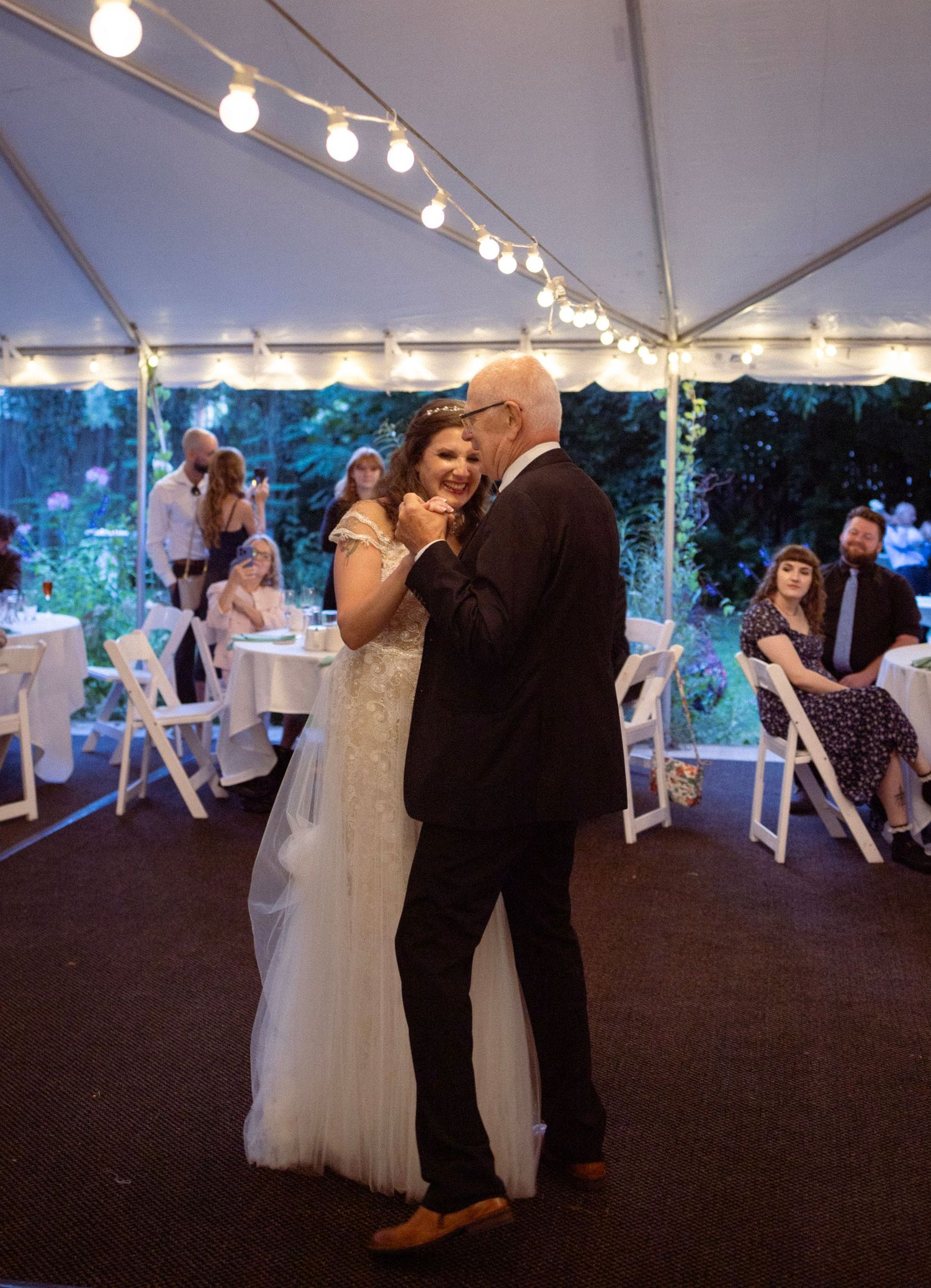 Bride and older man share a father-daughter dance under string lights in a white tent at an outdoor wedding reception.