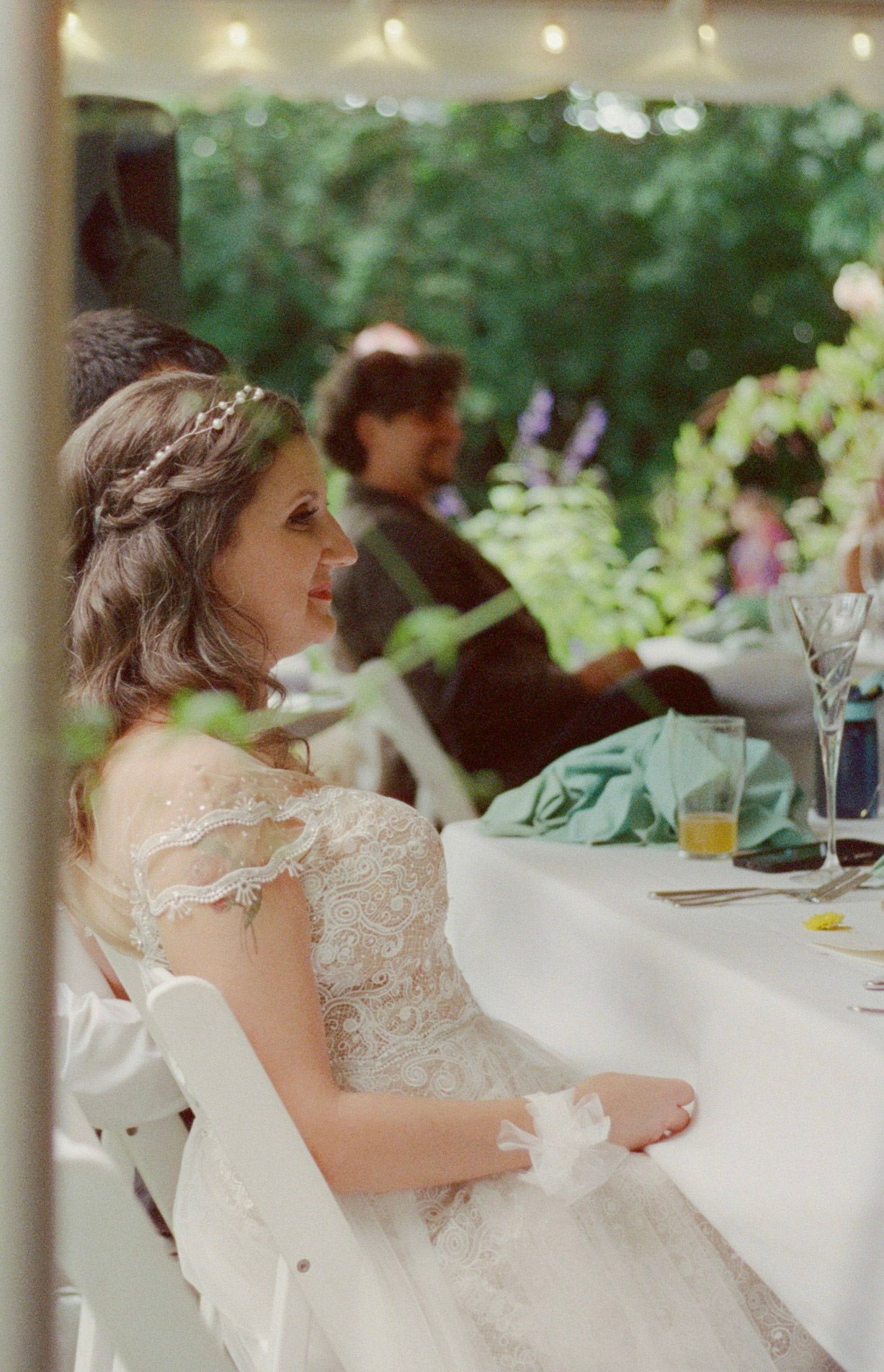 Bride in lace off-shoulder gown with floral crown sits at outdoor wedding reception table surrounded by greenery.