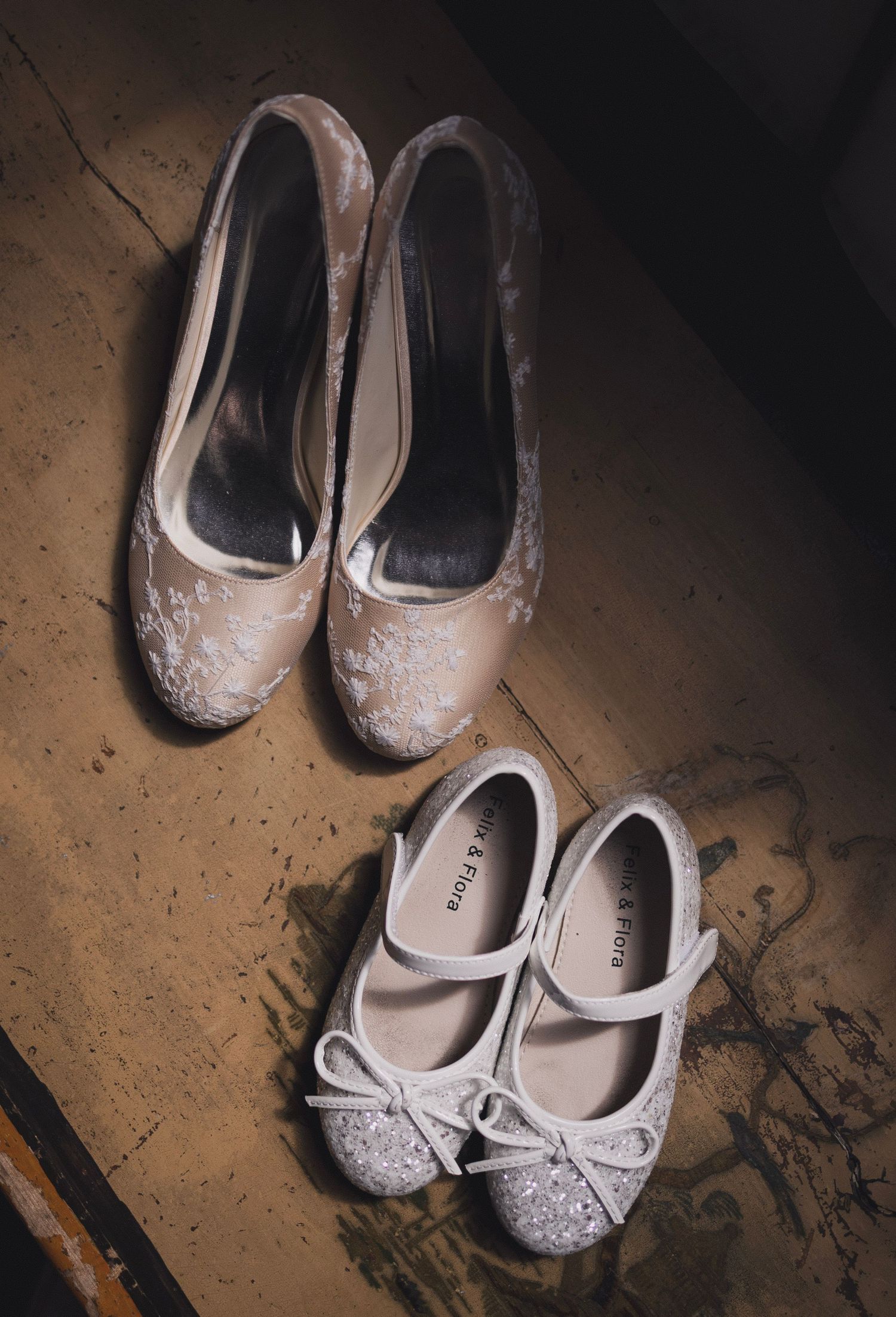 A pair of lace bridal heels and small girls glitter flats placed side by side on a wooden floor.