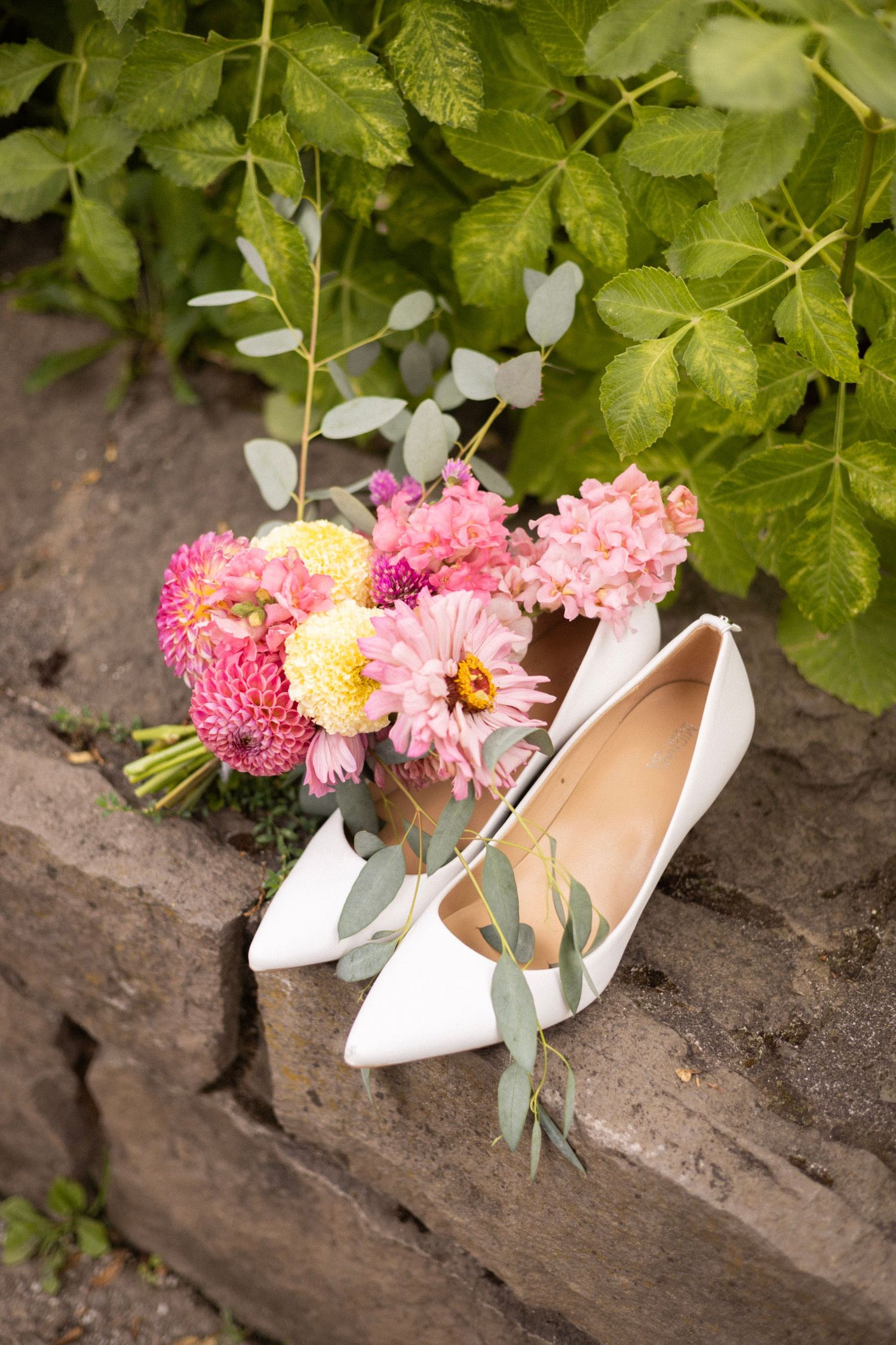 White bridal heels beside pink and yellow dahlia bouquet with eucalyptus on stone steps outdoors.