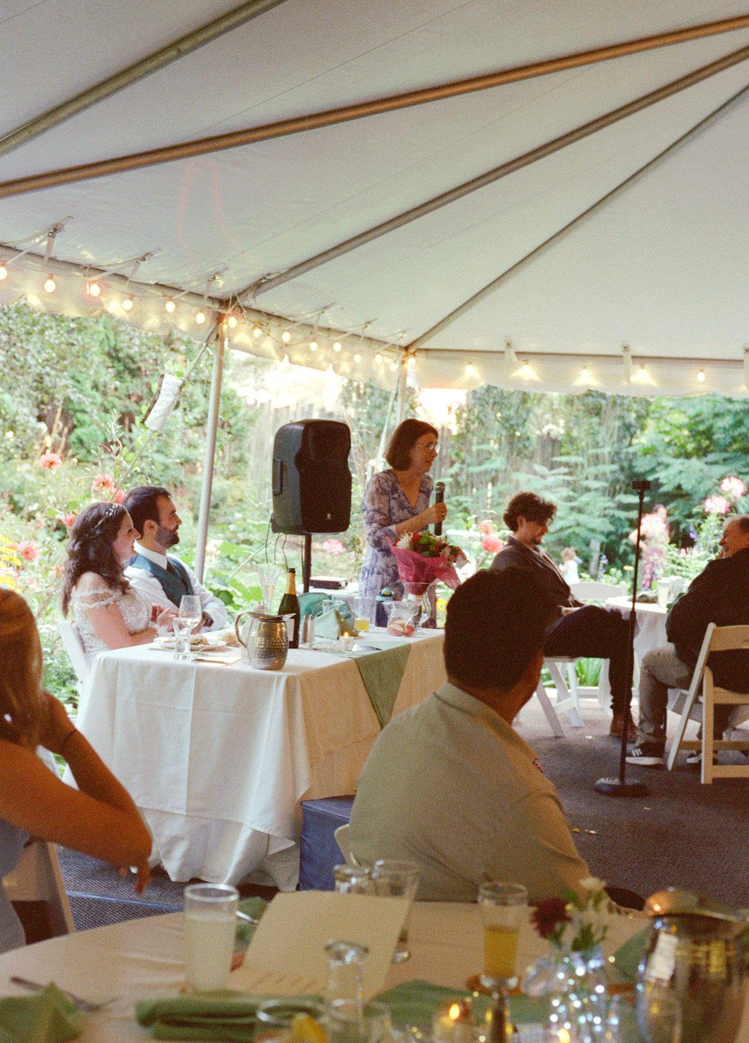 Outdoor wedding reception under a white tent with string lights, guests seated at tables while a woman gives a toast.