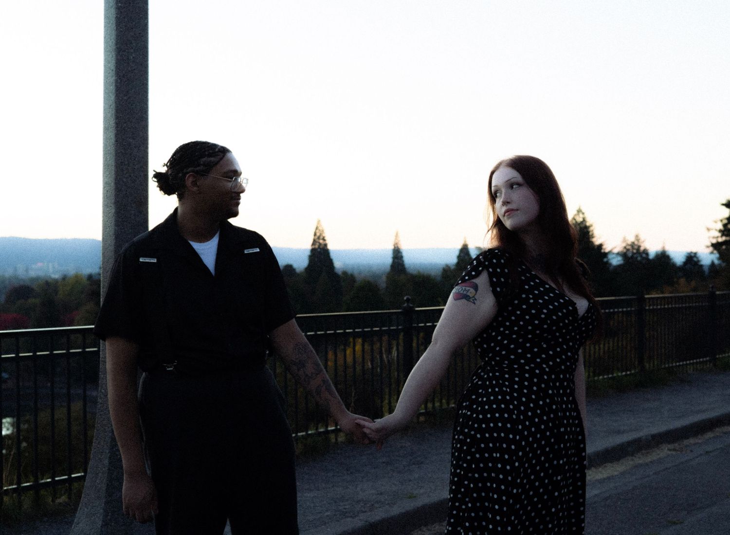 Couple holding hands outdoors at dusk with scenic tree-lined valley and tall monument in background.
