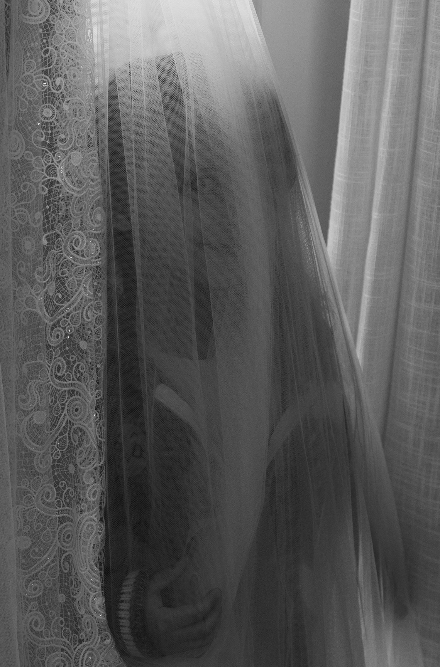 Black and white portrait of a woman partially hidden behind sheer lace curtains.