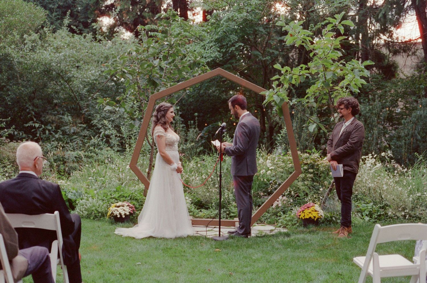 Outdoor wedding ceremony under hexagonal wooden arch in lush garden, bride and groom exchanging vows with officiant.