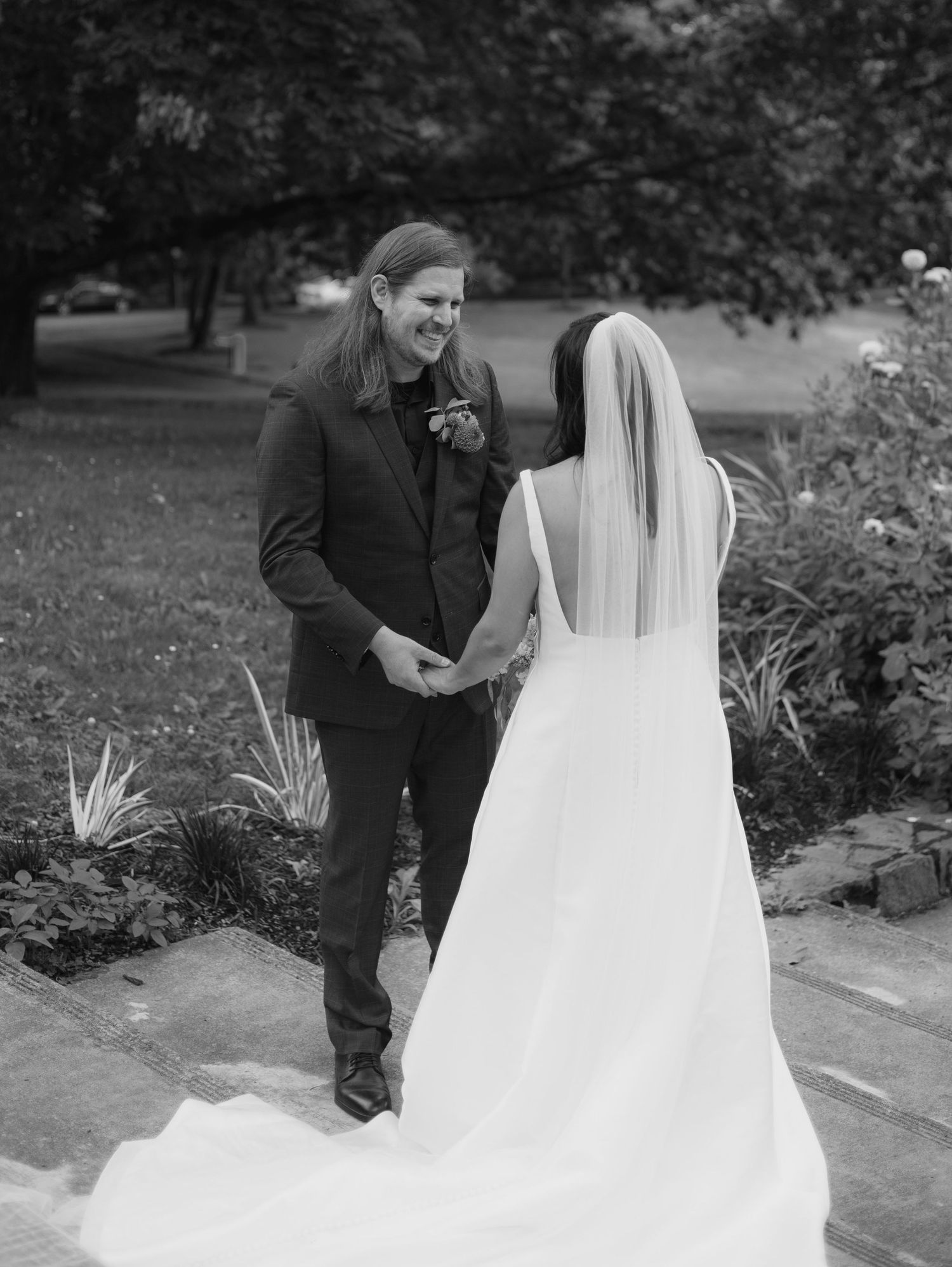 Black and white photo of a smiling groom holding hands with his bride in a wedding veil outdoors.