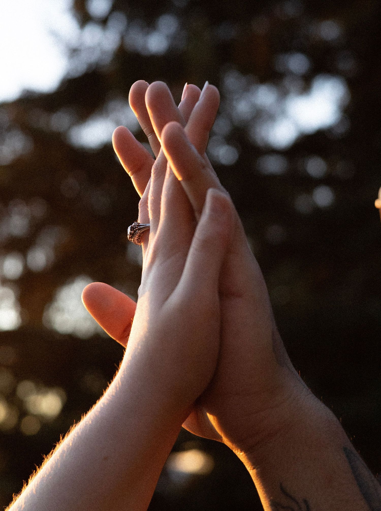 Two hands pressed together palm-to-palm outdoors at sunset, with a ring visible on one finger and a tattoo on a wrist.