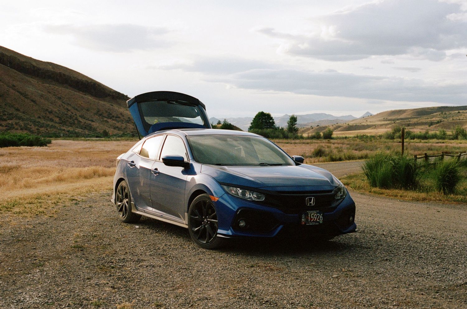 Blue Honda Civic hatchback parked on a gravel road with scenic rolling hills in the background.