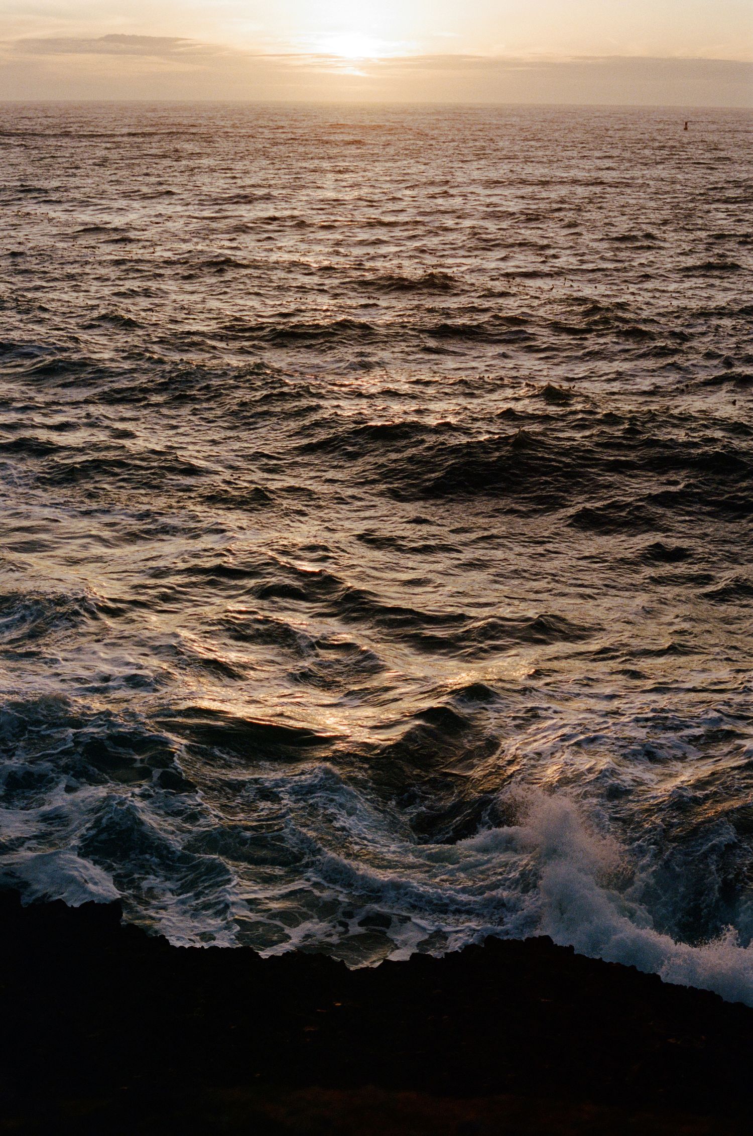 Dramatic ocean waves crash against rocks during a golden sunset over the open sea.