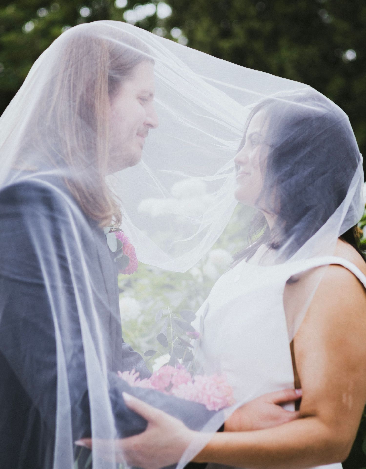 Bride and groom share romantic moment under flowing white wedding veil, holding pink and white floral bouquet outdoors.