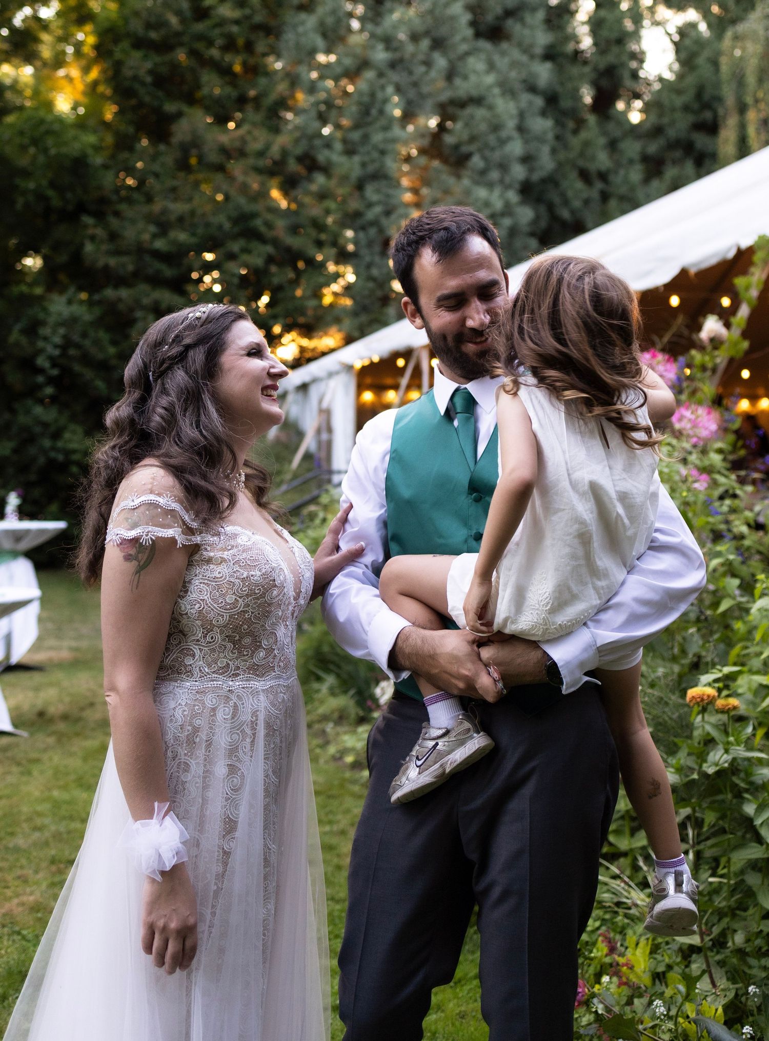 Bride and groom laughing with flower girl at outdoor garden wedding reception with string lights.