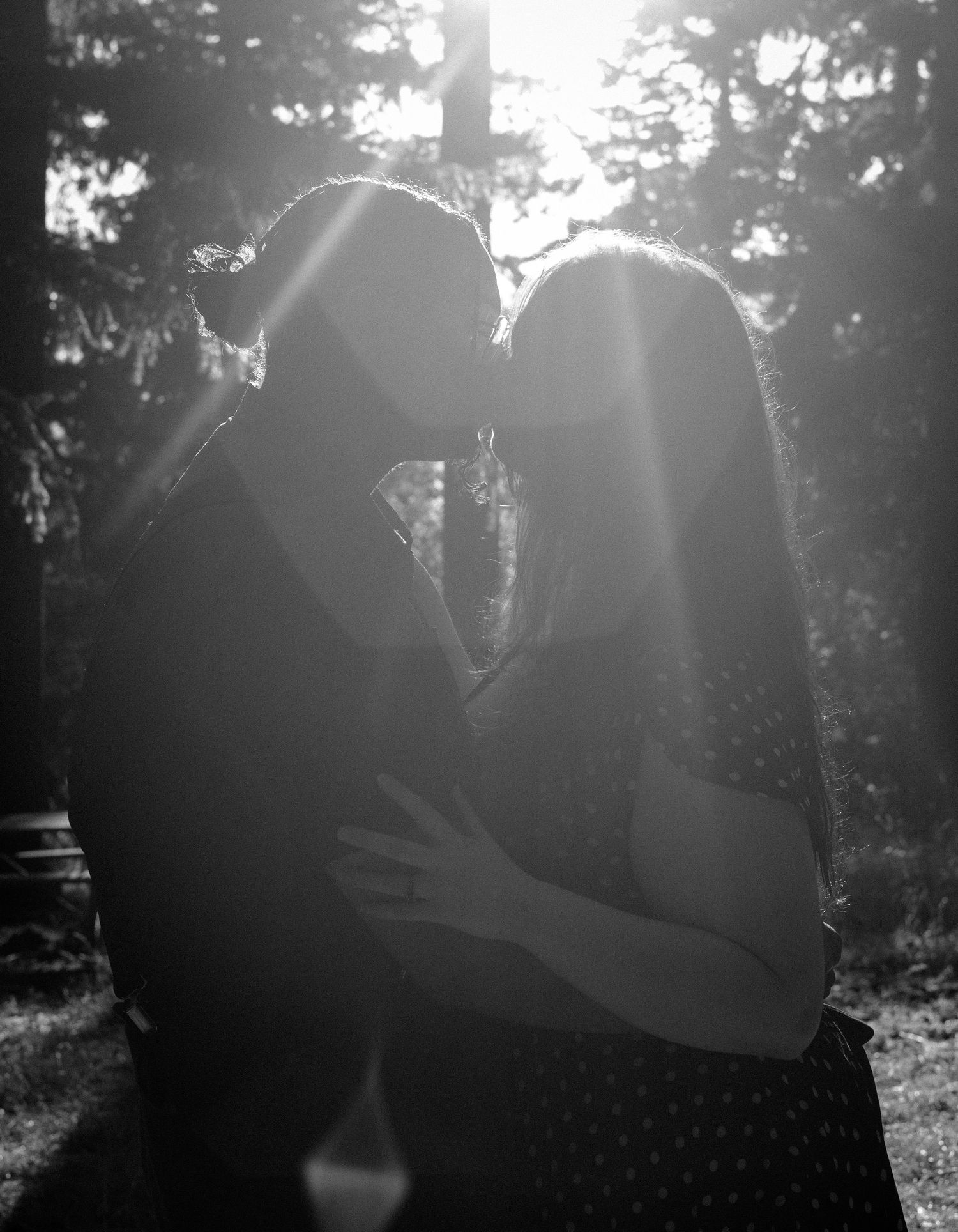 Black and white photo of a couple sharing a romantic kiss outdoors with sunlight streaming through trees.