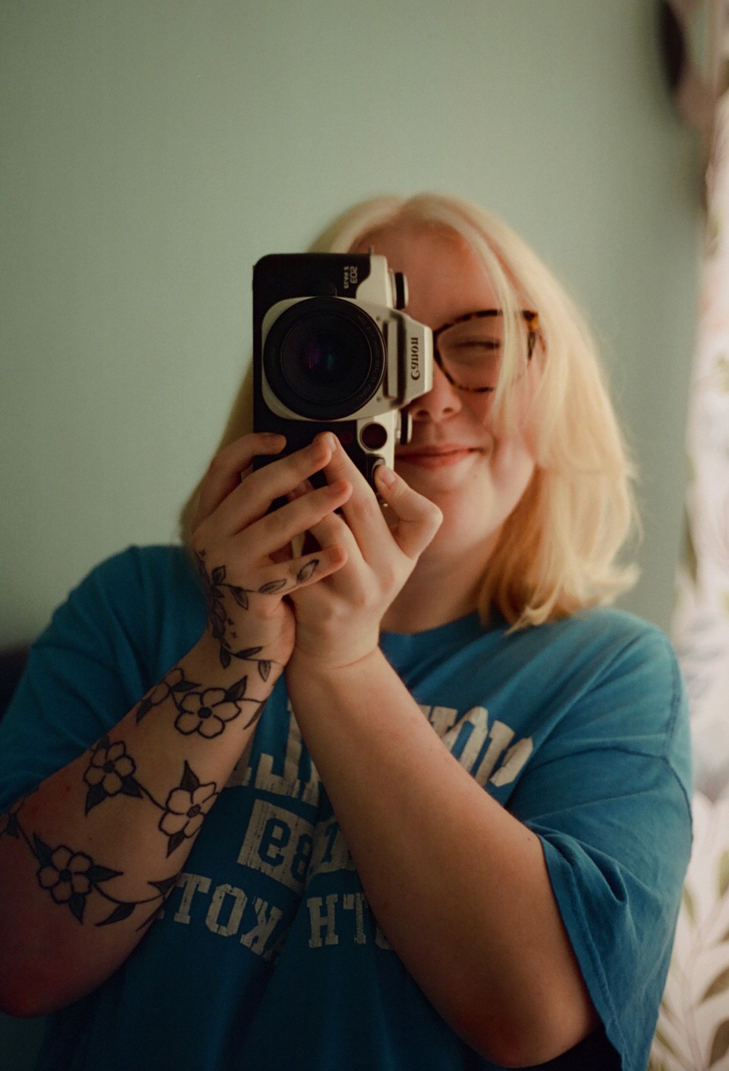 Tattooed blonde woman smiles while taking mirror selfie with Canon Elan ii camera.