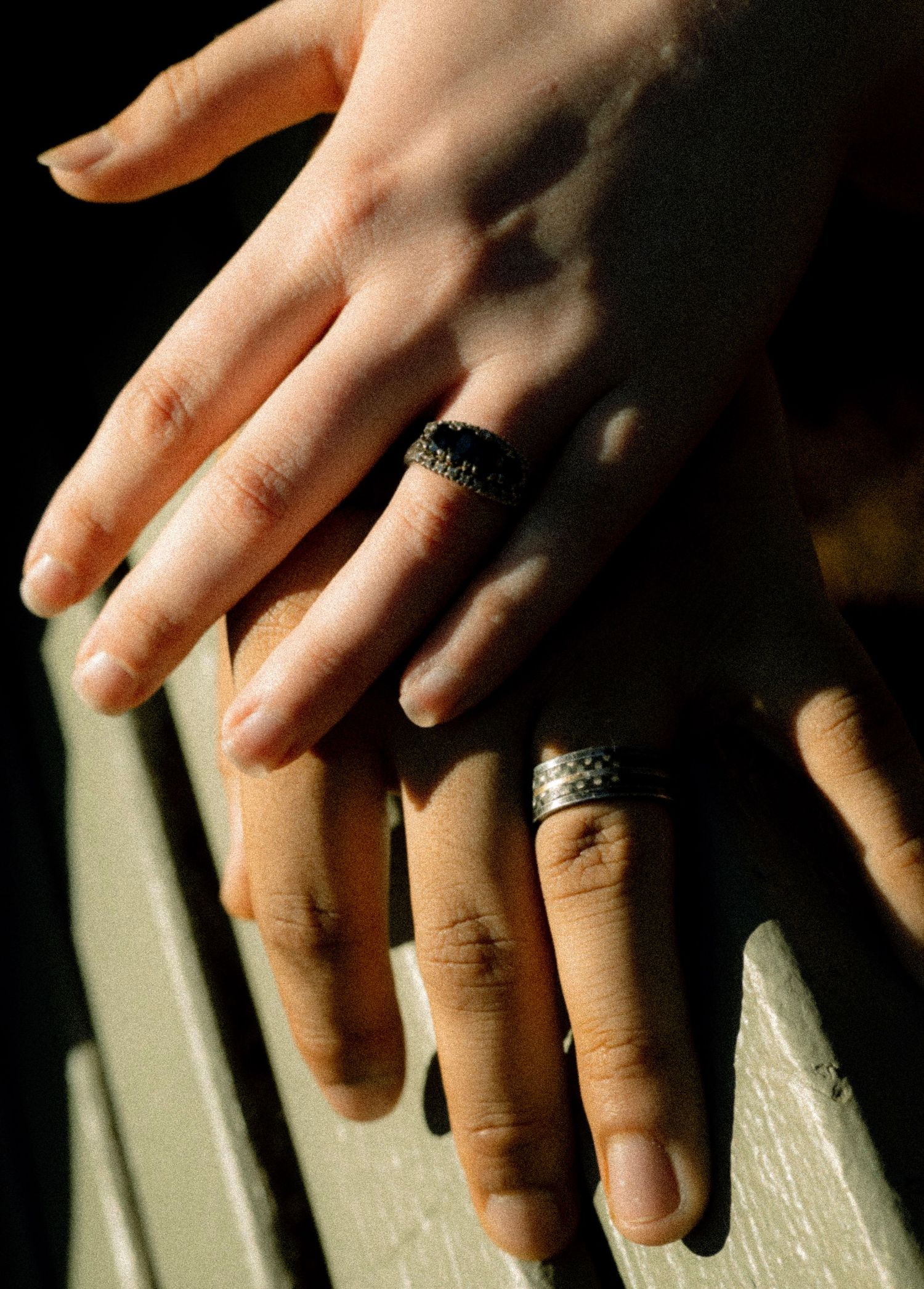 Two hands with rings, one lighter and one darker skin tone, resting together against a wooden surface.