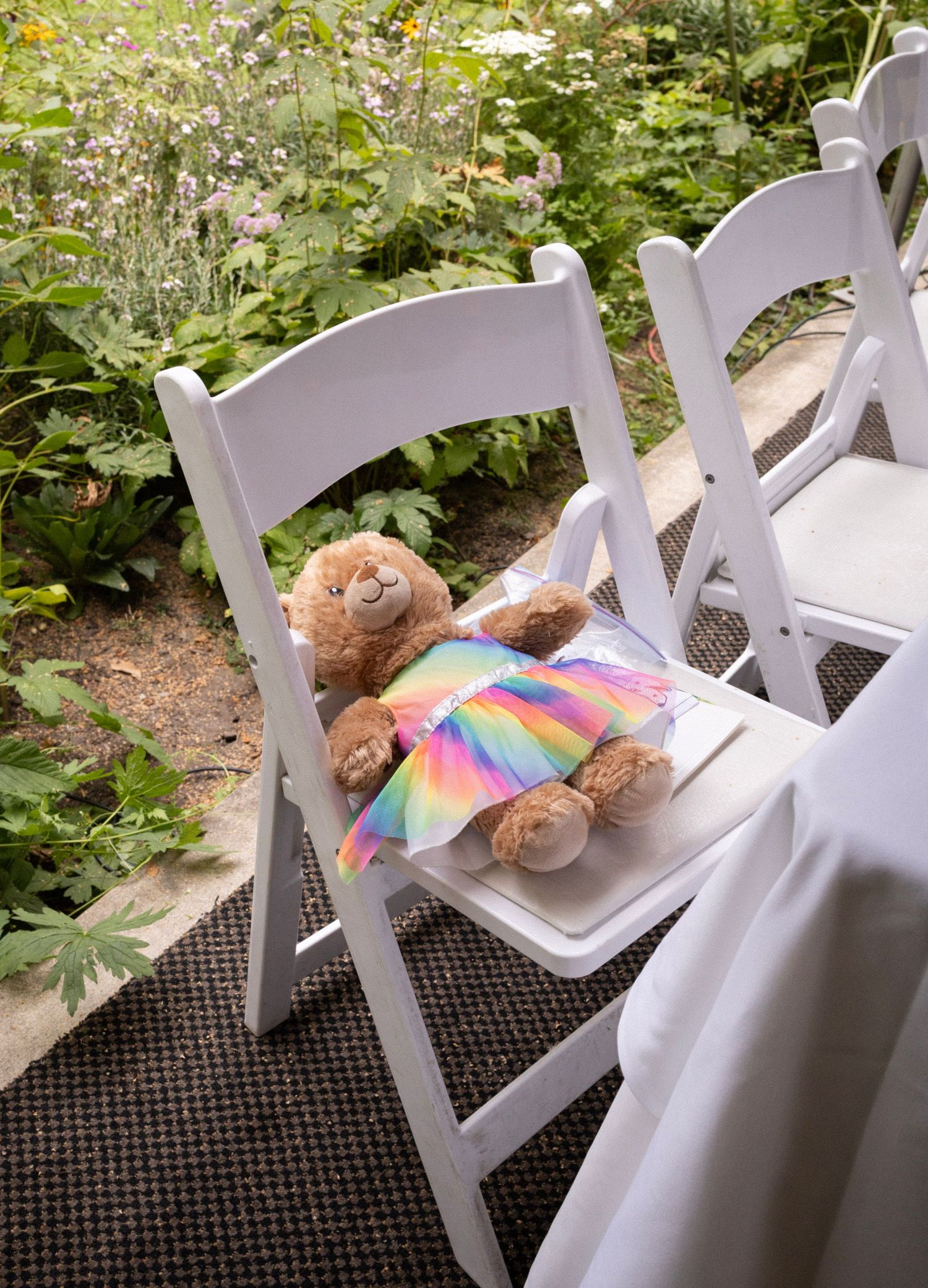 Adorable teddy bear in rainbow dress placed on white folding chair at outdoor garden wedding ceremony.