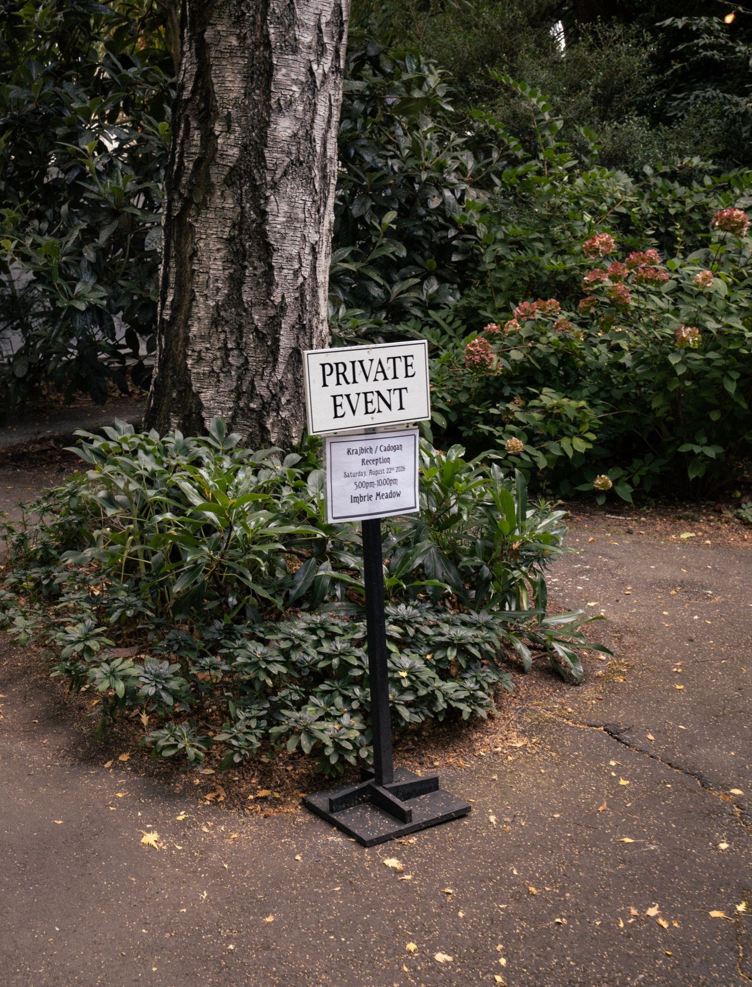 A private event sign on a black stand surrounded by lush green shrubs and trees in an outdoor garden setting.