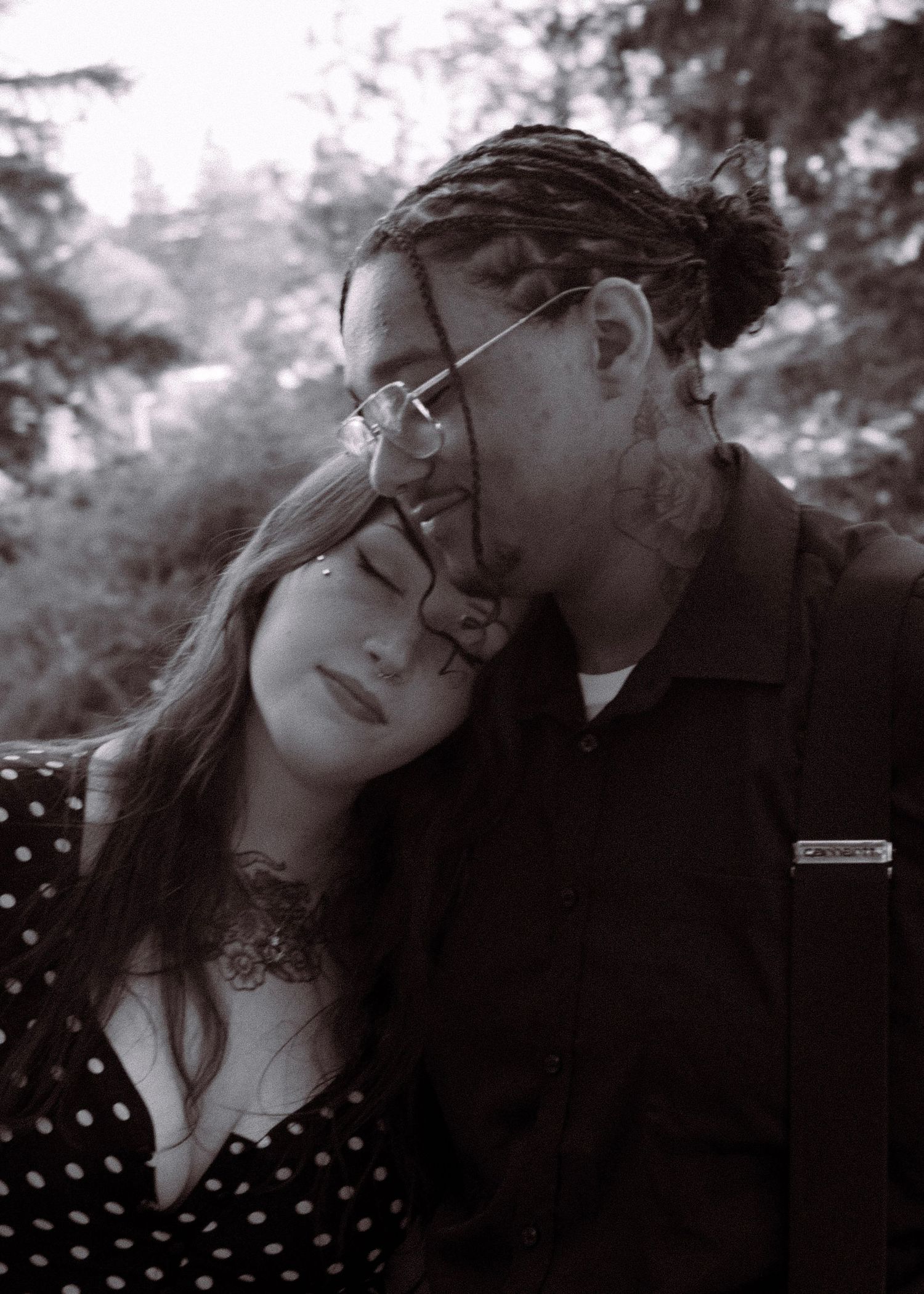 Black and white photo of a couple sharing a tender moment, woman resting head on mans shoulder outdoors.