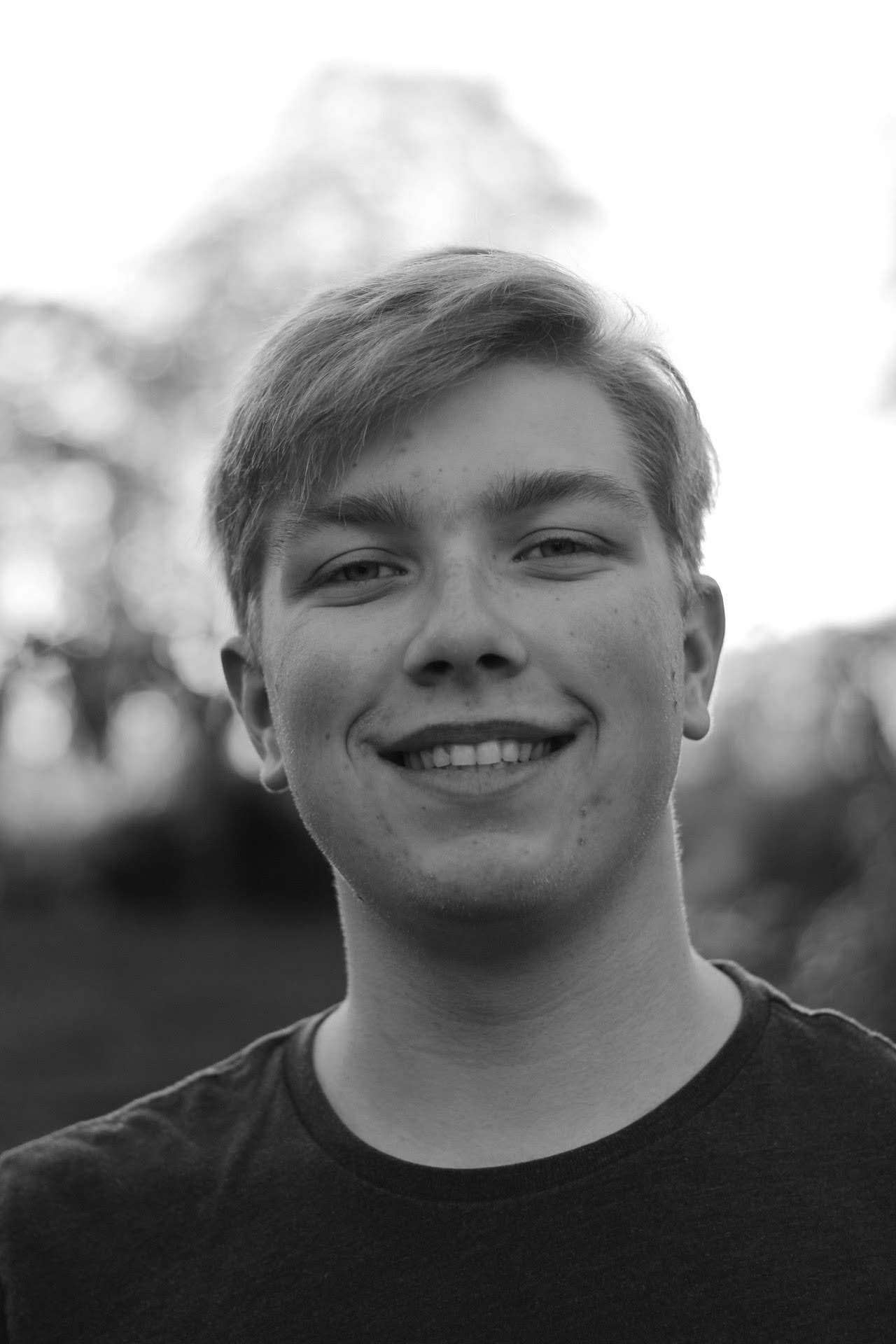 A smiling young man with blonde hair photographed in black and white outdoors.